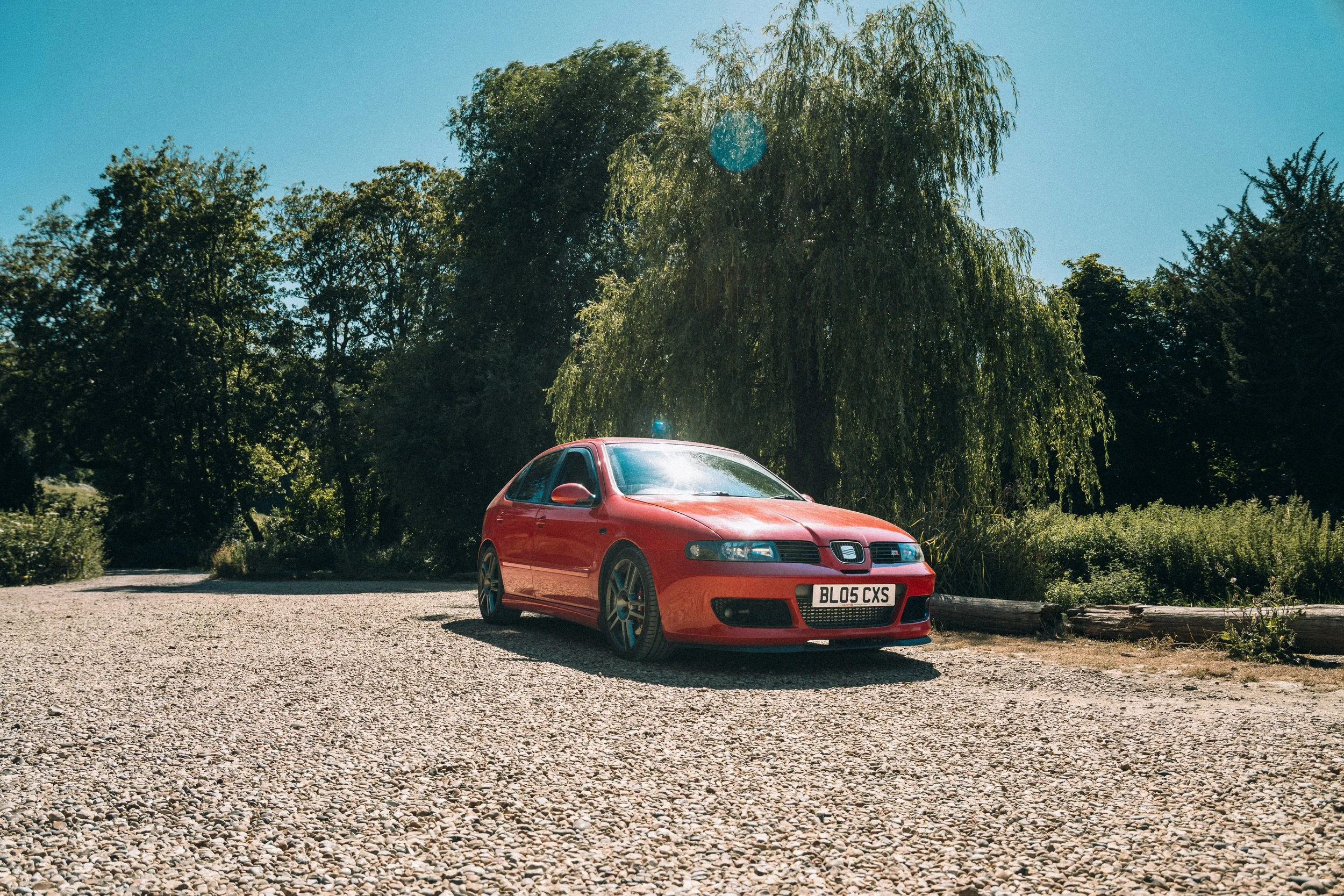 A red SEAT car parked on a gravel surface with a lush green background of trees and a clear blue sky.