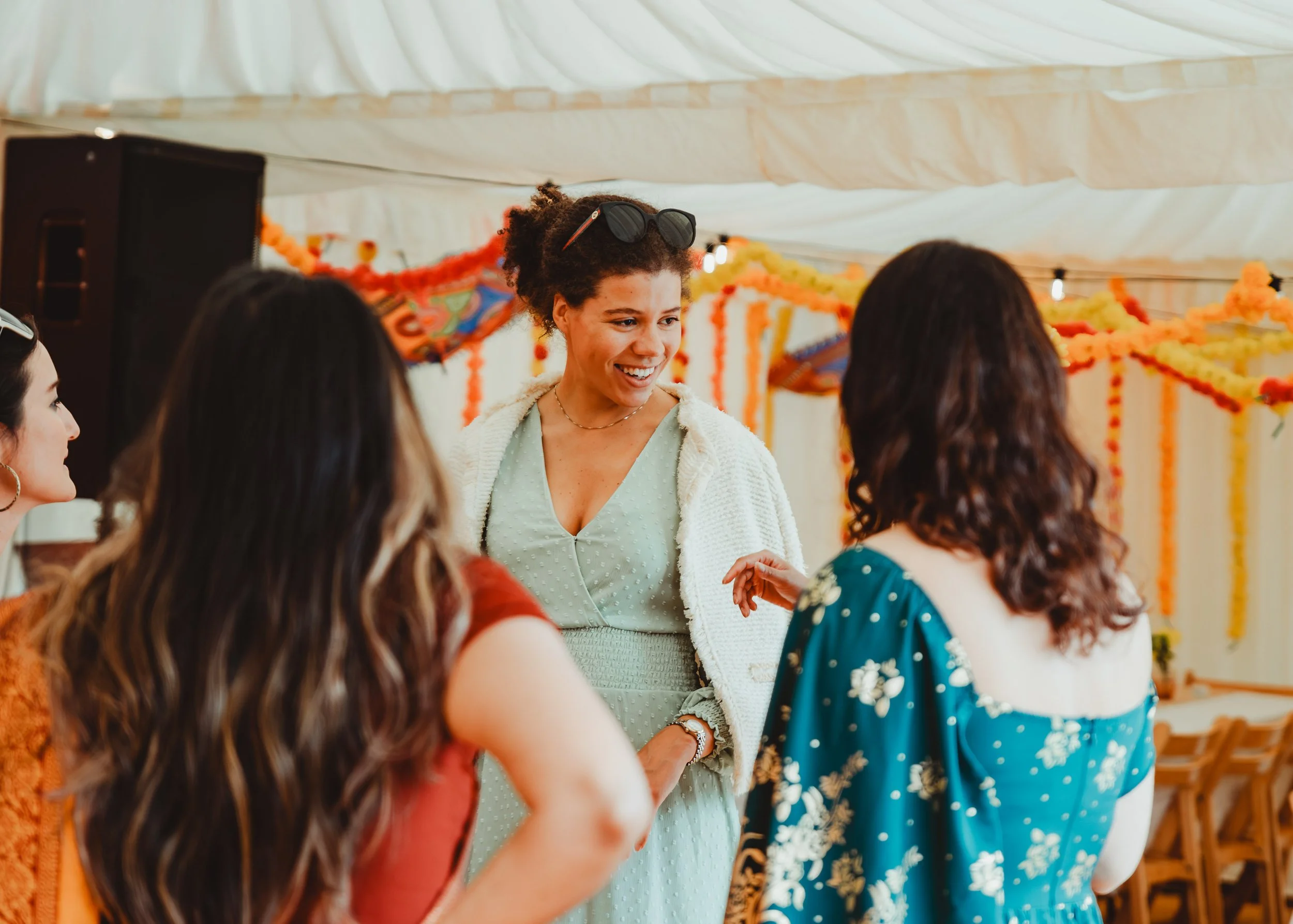 A group of women gathered under a decorated tent, engaging in conversation, with a woman smiling in the center, wearing sunglasses on her head.