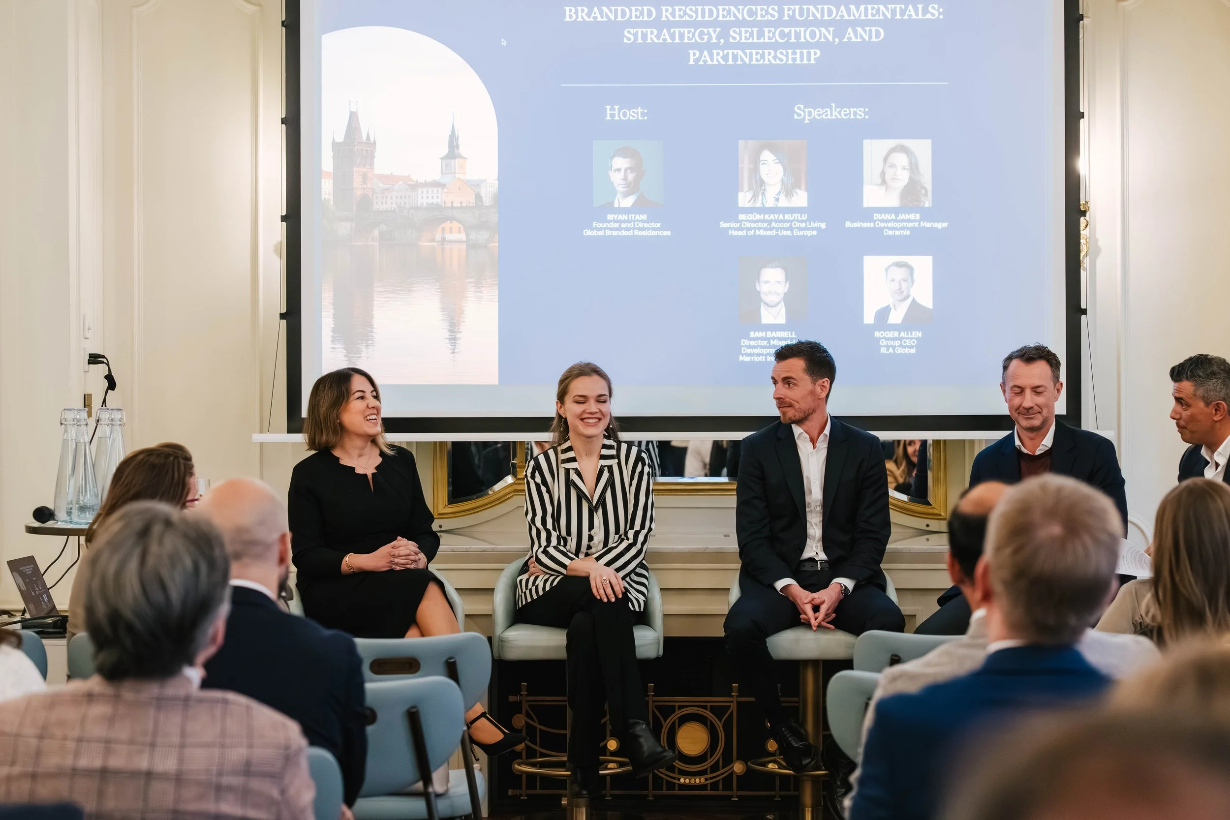 A group of people seated in a conference room, listening to a panel discussion. Four panelists are seated in front of a large screen displaying a presentation titled 'Branded Residences Fundamentals: Strategy, Selection, and Partnership.' The panelis