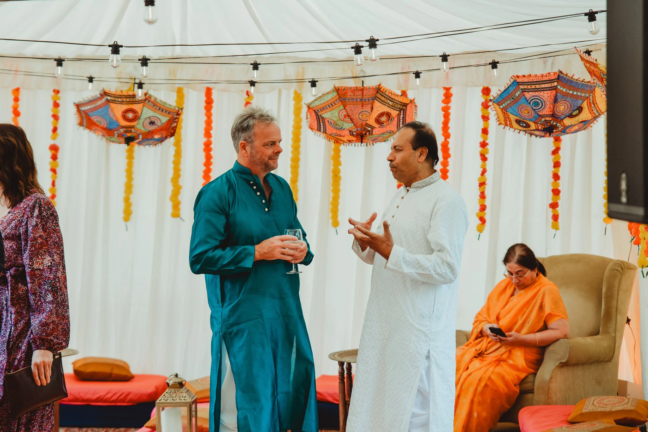 Two men dressed in traditional Indian attire are conversing, one holding a glass of wine, inside a decorated hall with umbrellas and marigold flower garlands hanging from the ceiling.