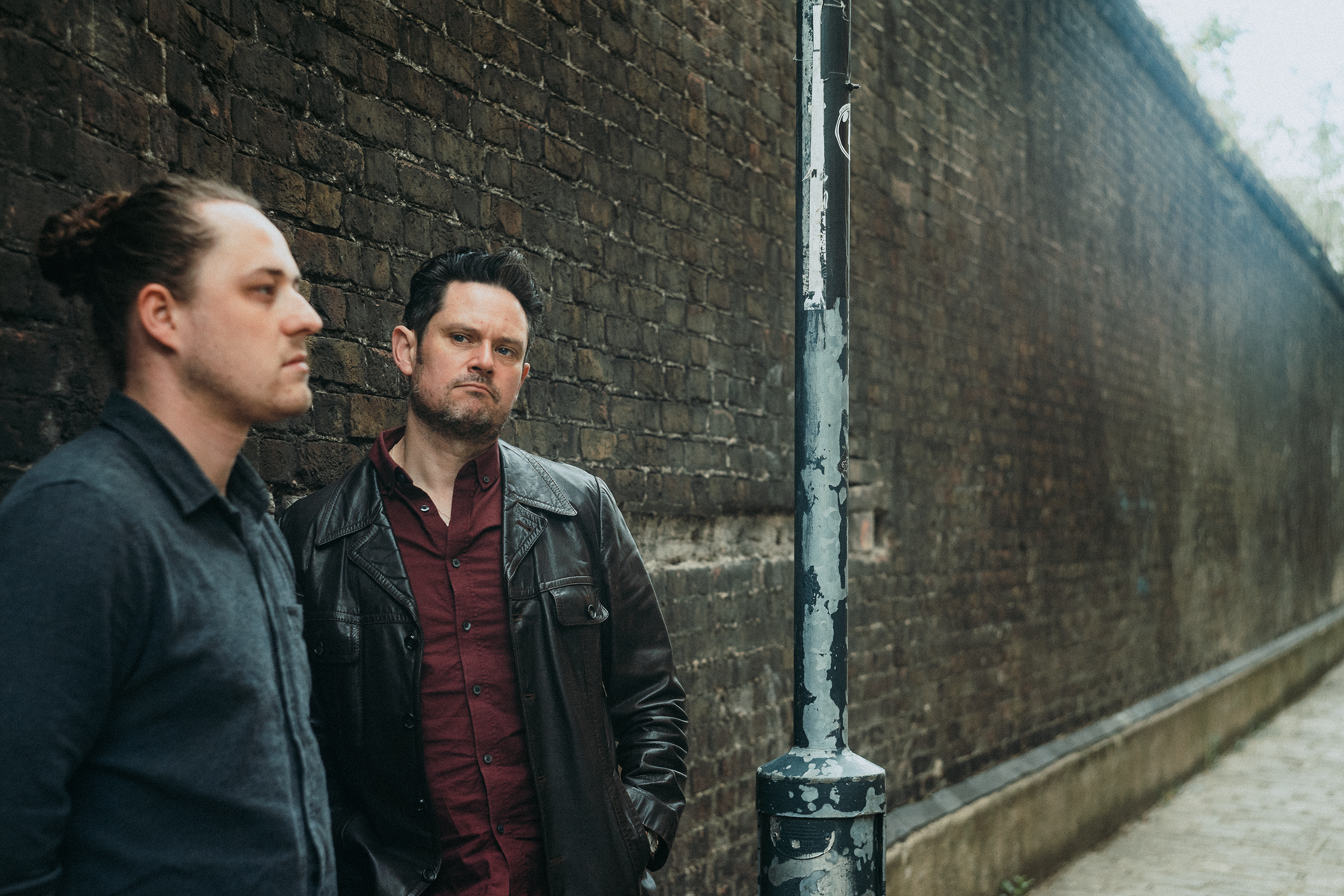 Two men with dark hair and serious expressions leaning against a brick wall outdoors near a weathered lamppost, in an urban setting.
