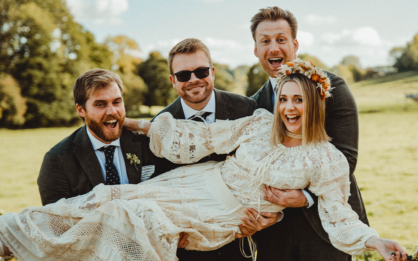 A woman in a vintage white lace dress and floral crown being held by four smiling men outdoors in a park