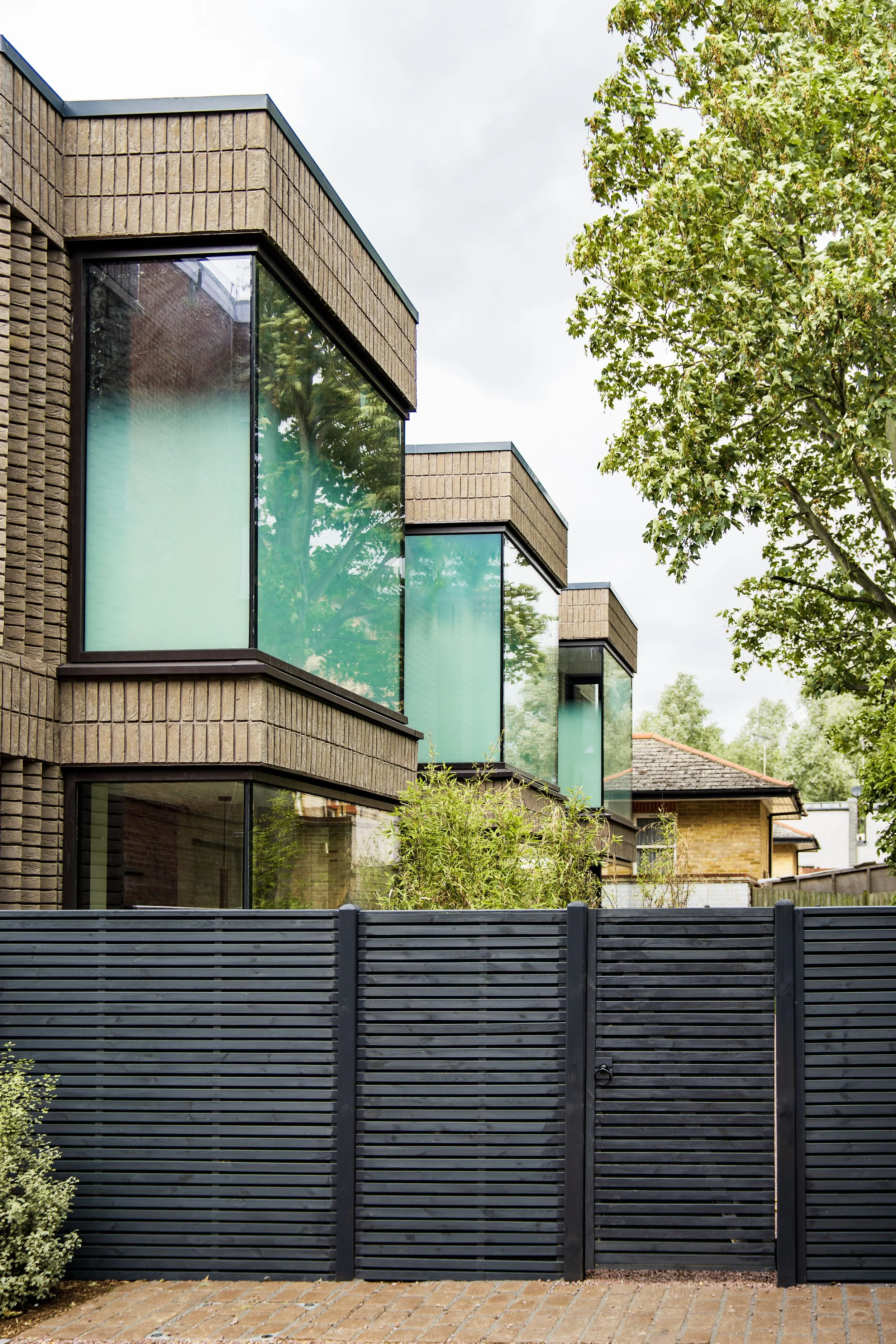 Modern brick building with large glass windows, black metal fence, and greenery in front.