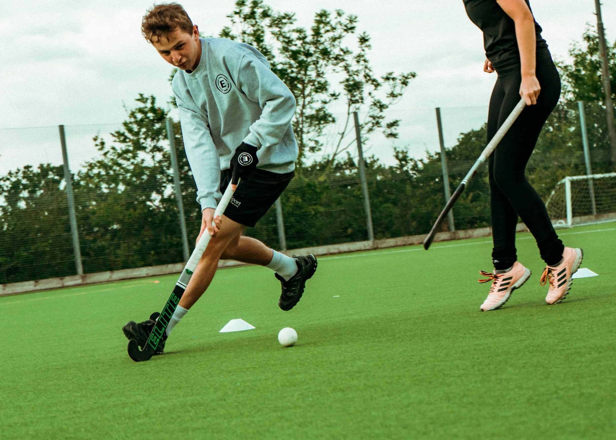Two people playing field hockey on a green outdoor turf, with one person bending down to hit the ball with a stick and the other person preparing for their turn, surrounded by trees and a fence.