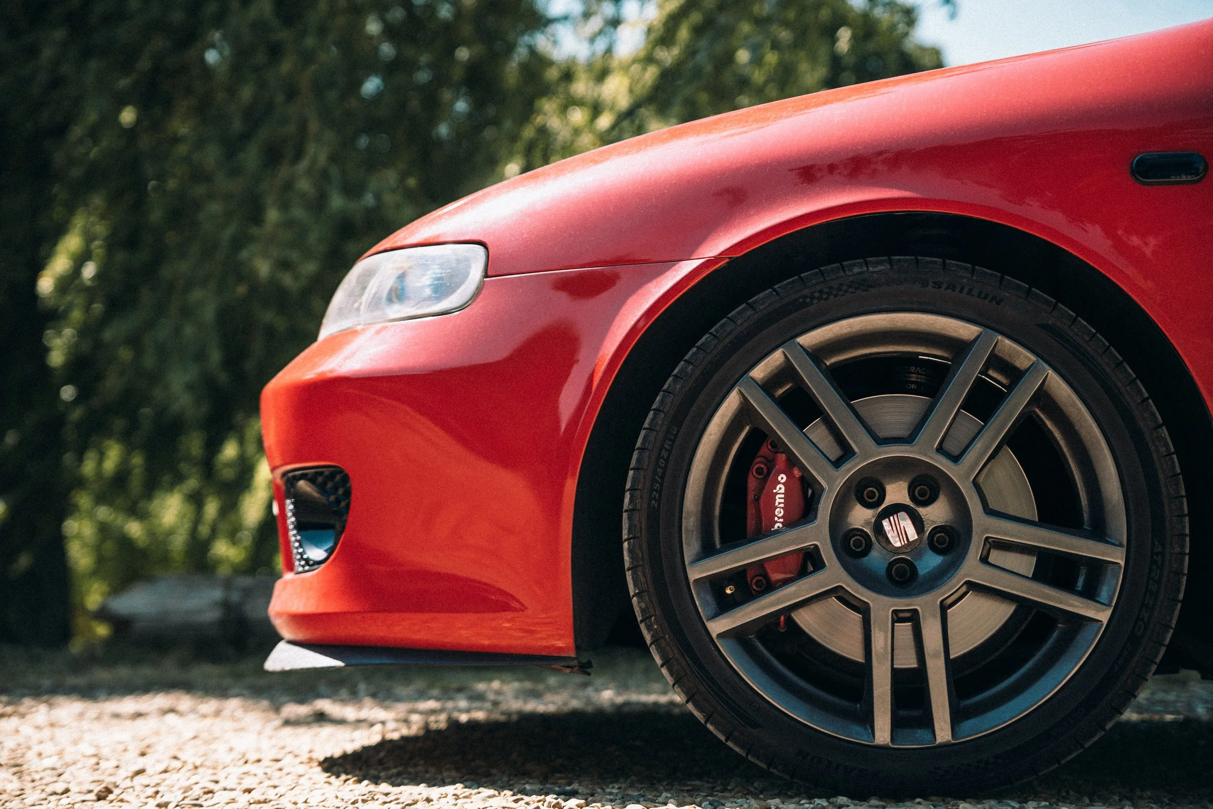 Close-up of the front left side of a red sports car, showing the wheel, headlight, and part of the fender, with a blurred outdoor background of green trees.
