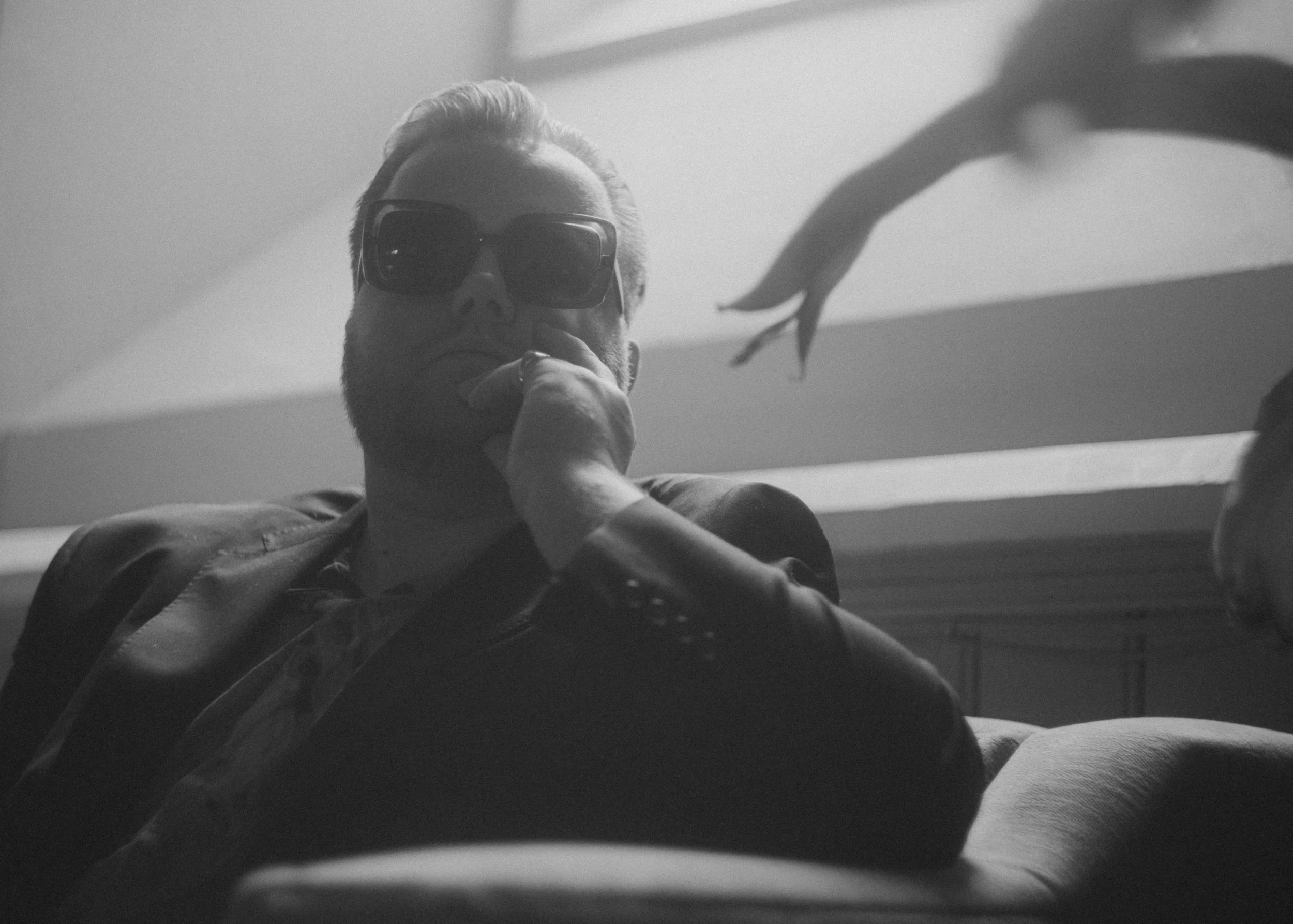 A man with a beard wearing sunglasses and a leather jacket sitting indoors, resting his chin on his hand, with a blurred human hand reaching towards him in the background, in black-and-white.