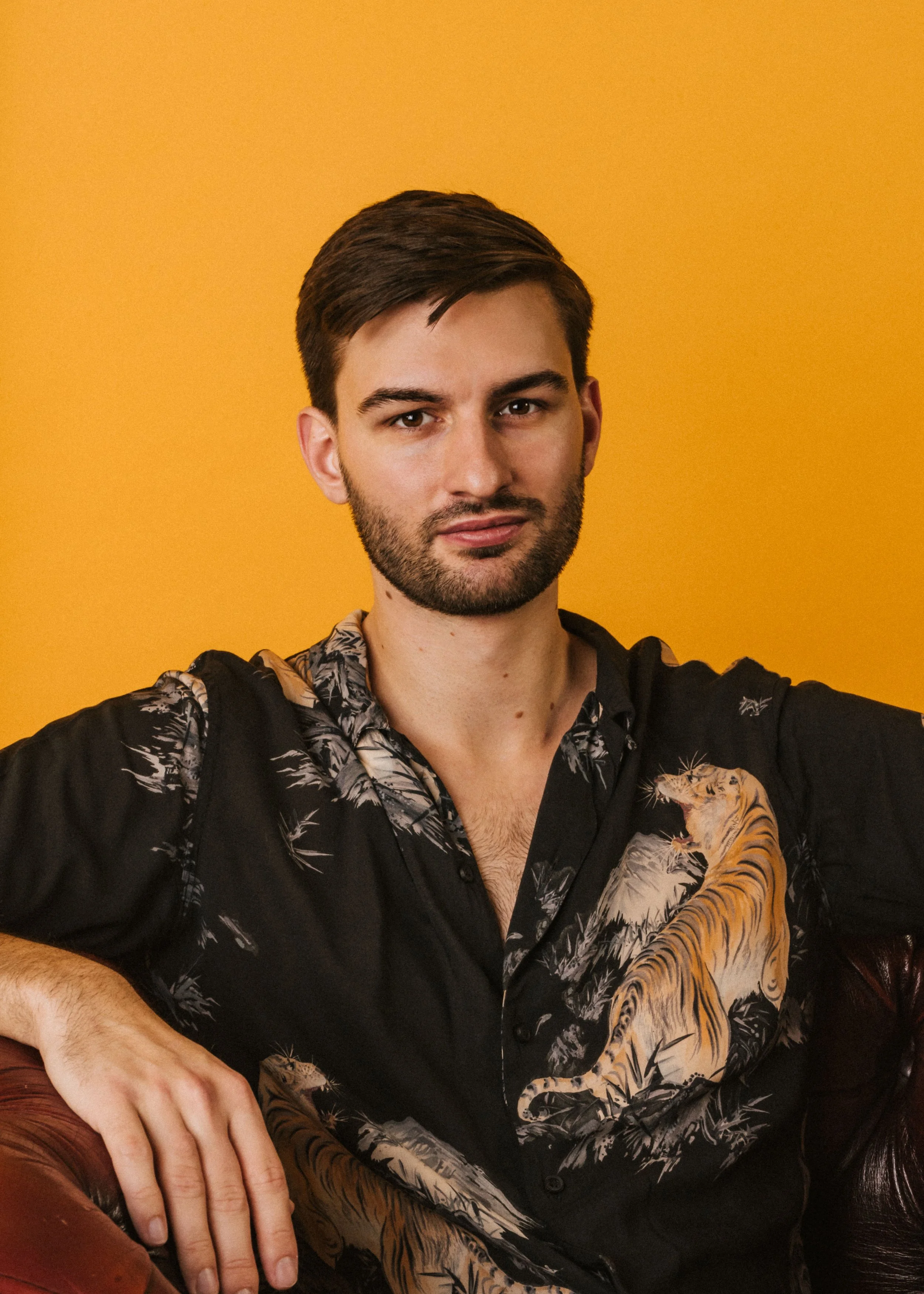 A young man with short dark hair and a beard, wearing a black shirt with a tiger design, sitting against a yellow background.