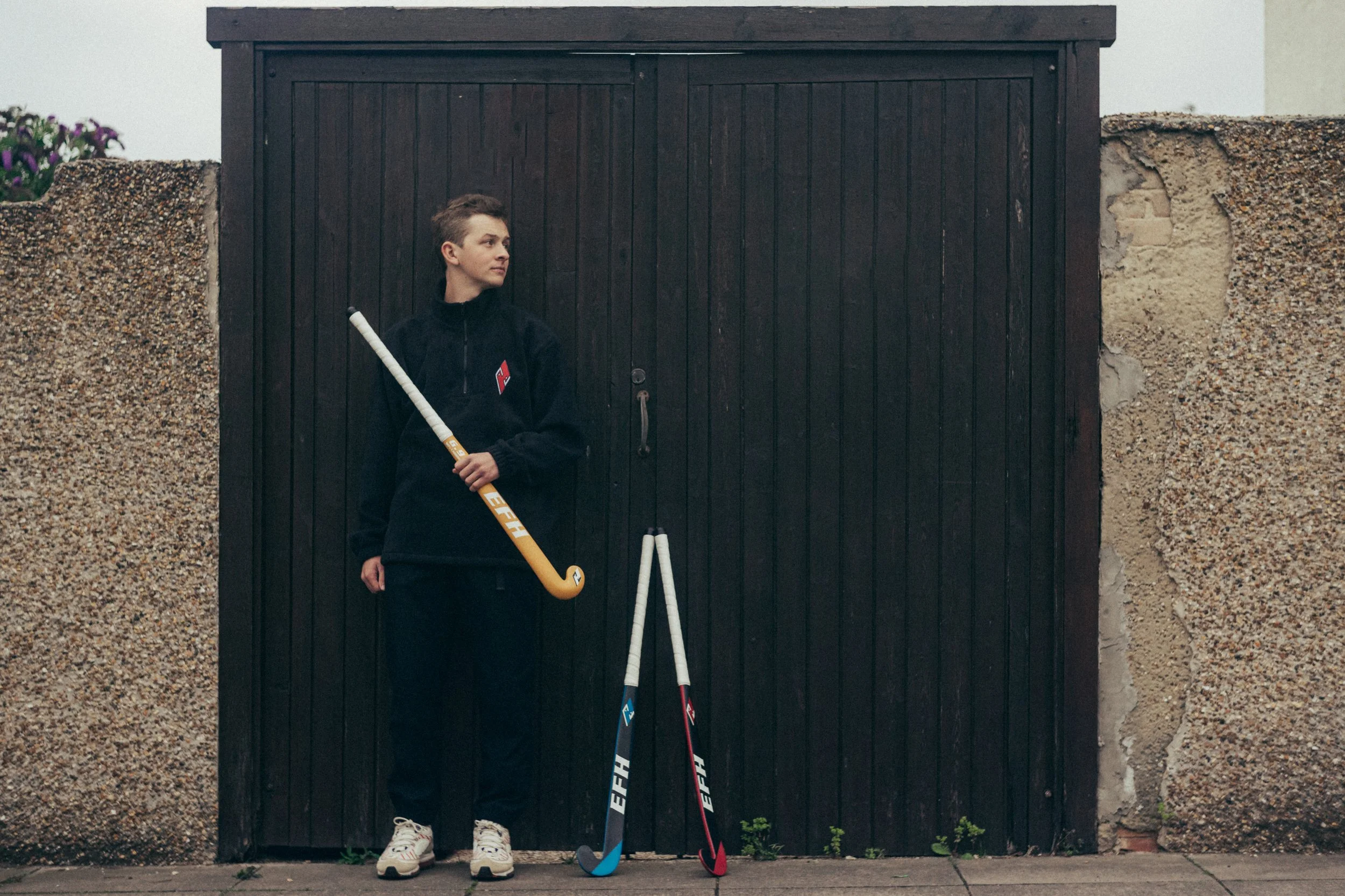 A young woman with short hair standing in front of a black wooden gate, holding a hockey stick. There are two additional hockey sticks leaning against the gate, and a wall with textured concrete on each side. She is wearing a black jacket, black pant