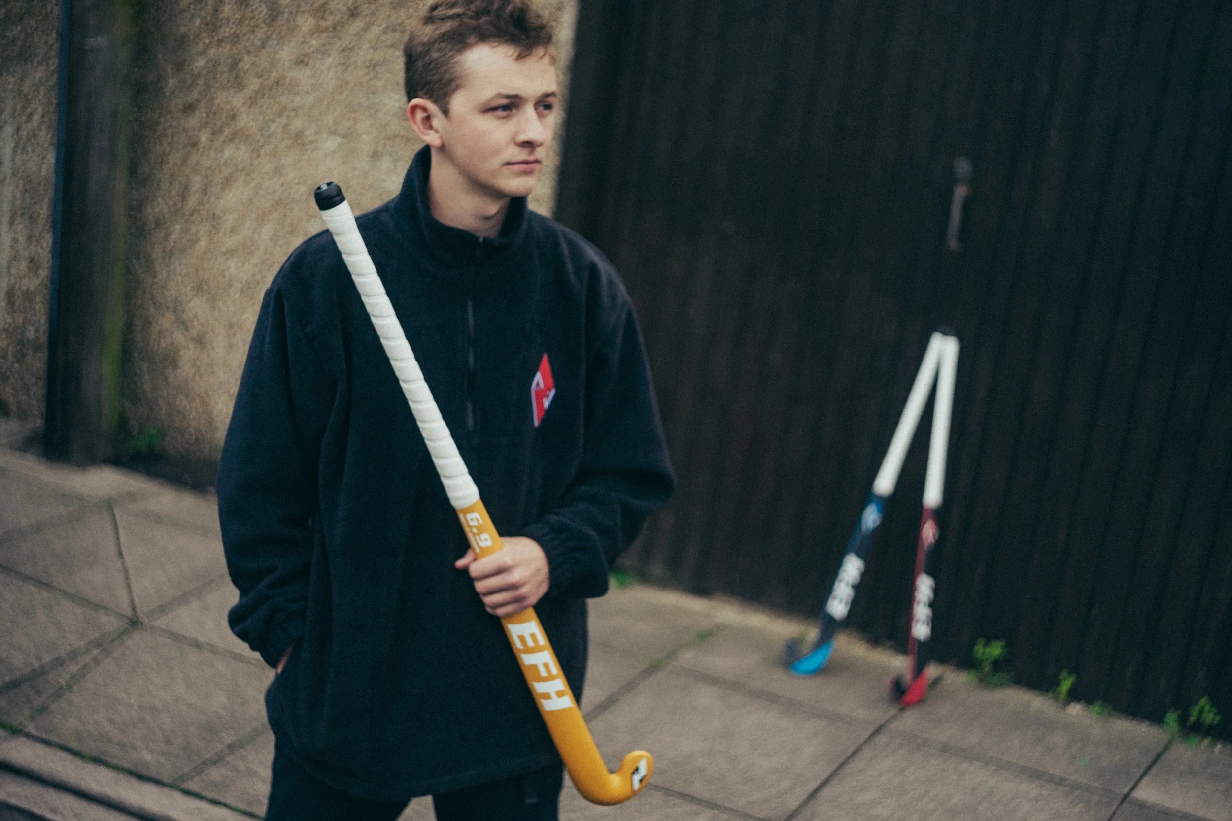 A young man standing outdoors on a sidewalk, holding a yellow hockey stick, with a serious expression, wearing a dark hoodie, near a dark fence and hockey equipment in the background.