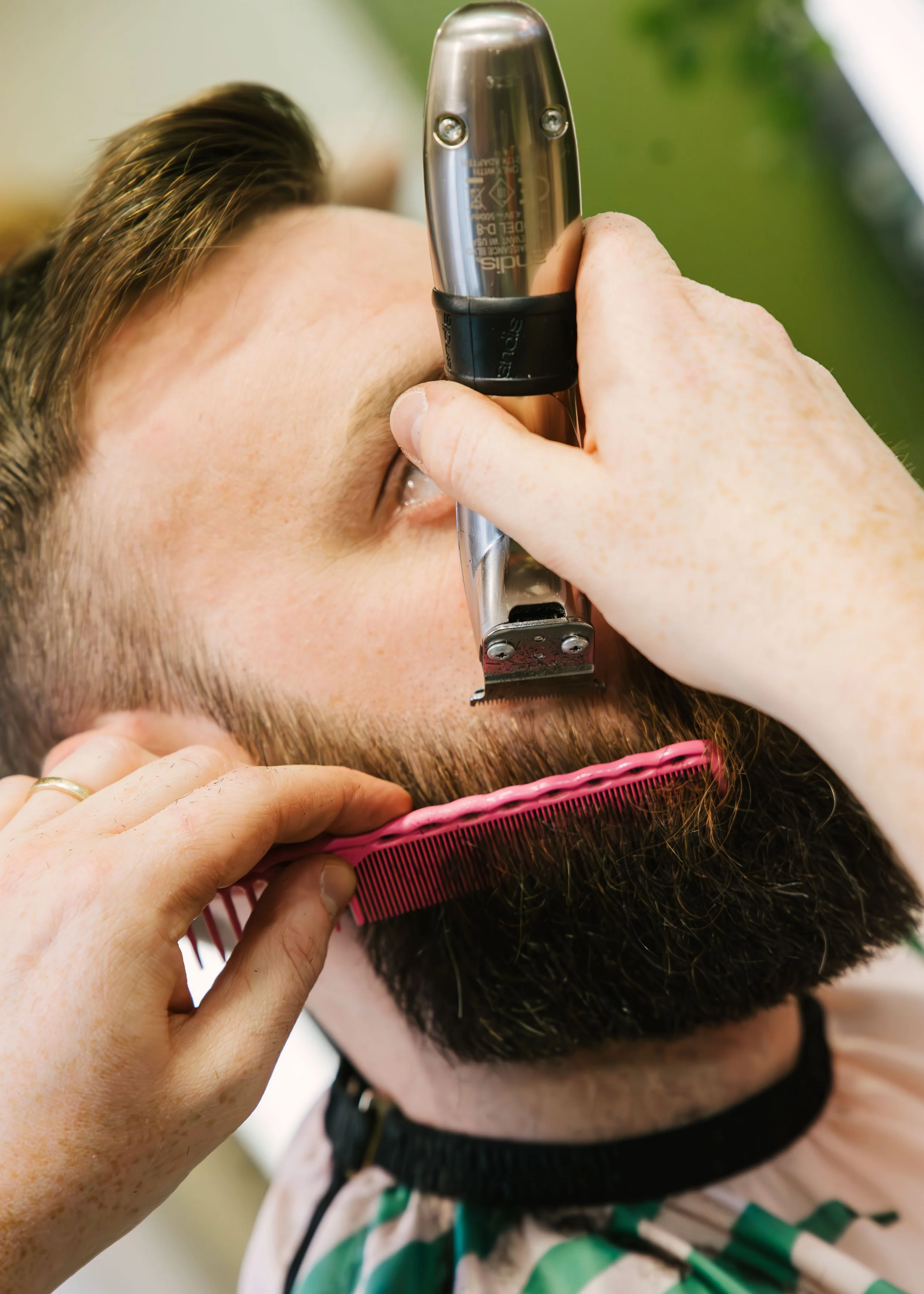Barber trimming a man's beard with electric trimmer, close-up of hand and hairdresser's tools.