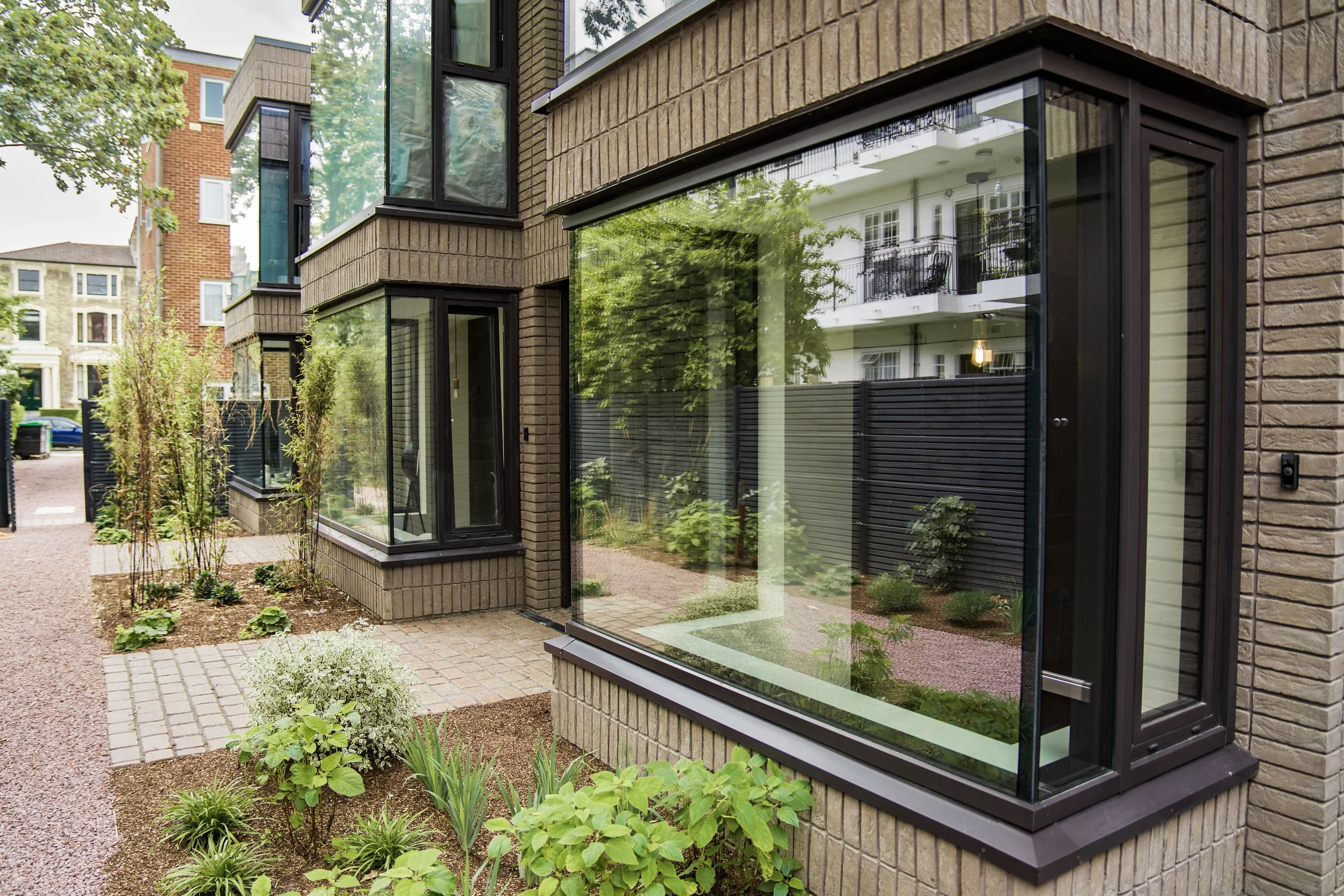 Exterior view of a modern building with large glass windows and brick walls, surrounded by a small garden with various plants and a gravel pathway.