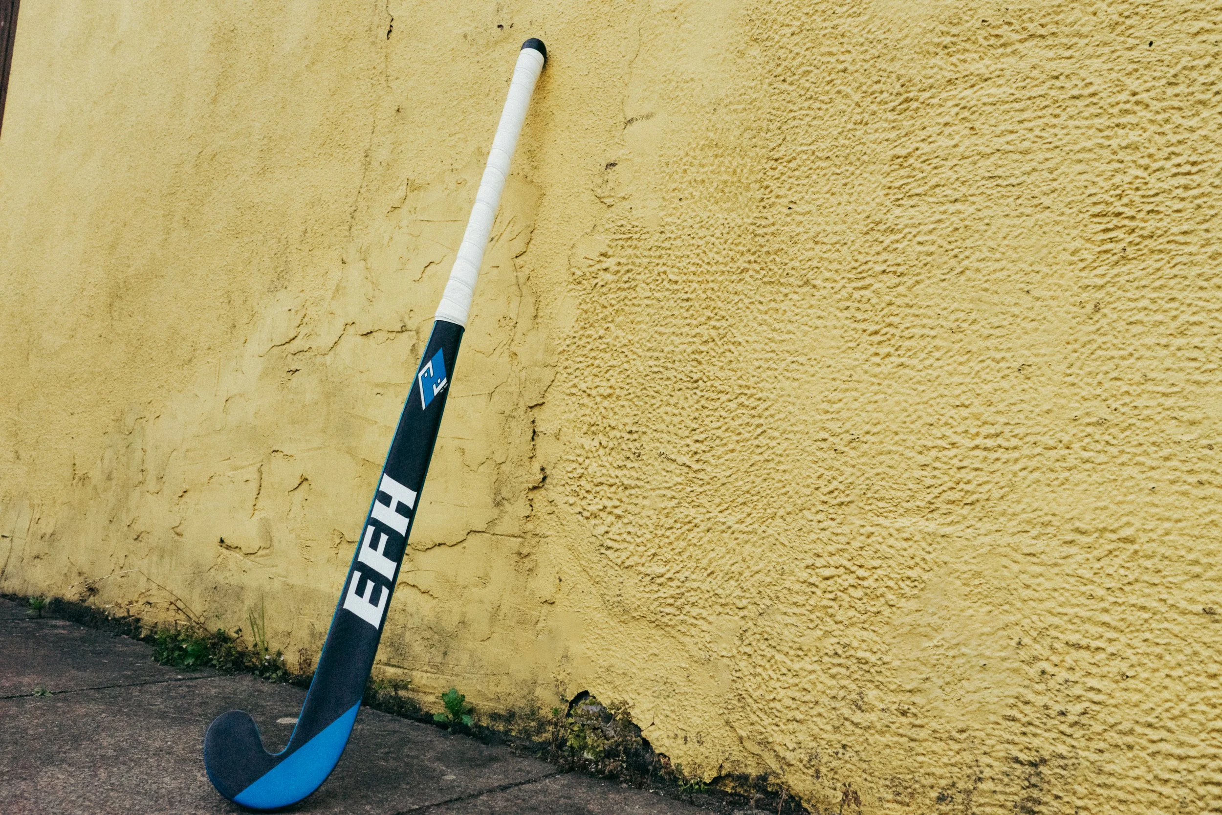 A blue and black hockey stick resting against a yellow textured wall outside, near the sidewalk.