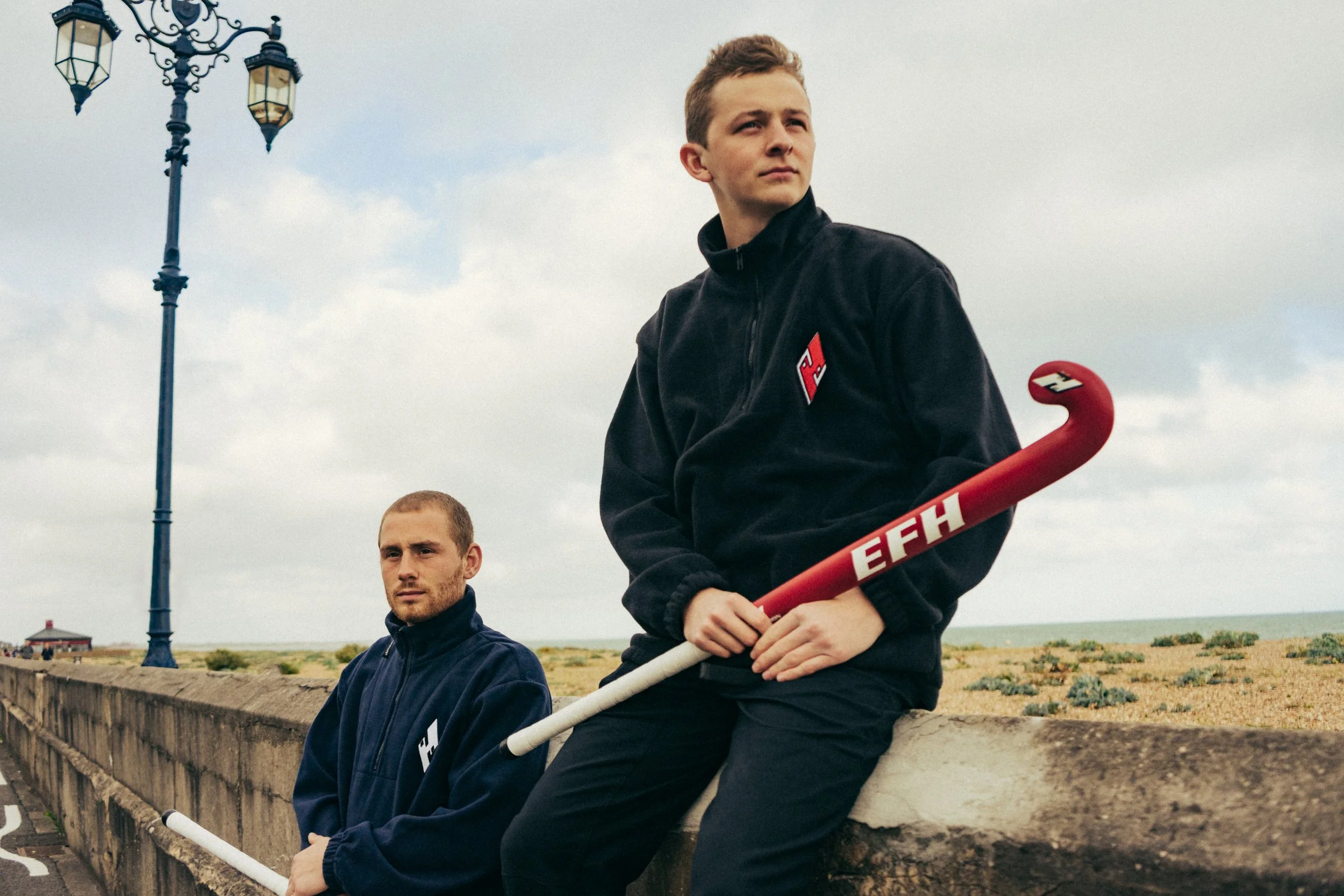 Two young men outdoors, one holding a red and white hockey stick, sitting near a concrete wall along a beach with grass and sand. A lamppost and a small building are visible in the background, with overcast skies.