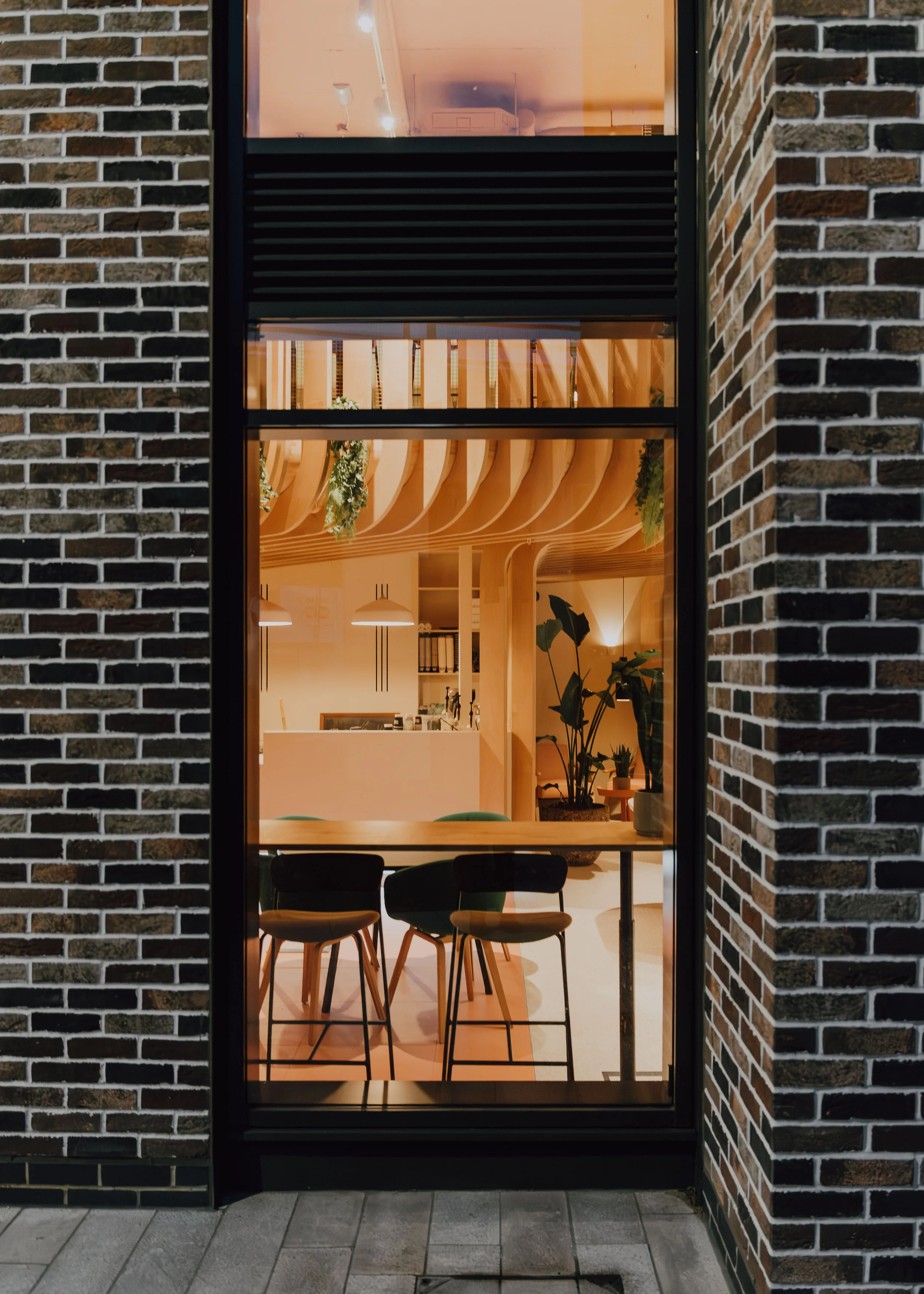 Interior of a modern cafe or restaurant with a high wooden table, black chairs, hanging pendant lighting, and indoor plants, seen through a large window by a brick wall.