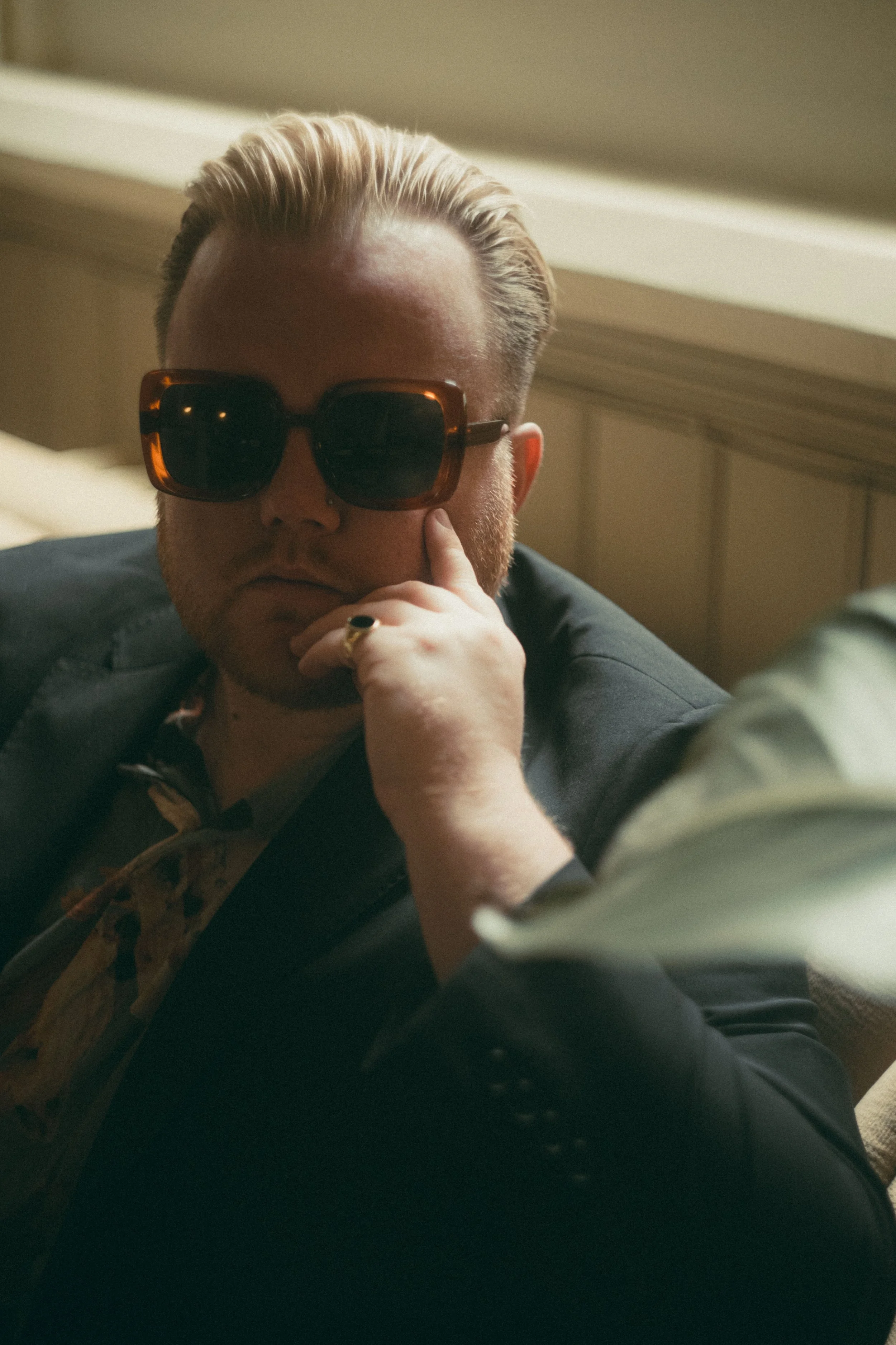 A person with blonde hair wearing dark sunglasses and a black jacket. They are resting their chin on their hand and appear to be indoors, possibly in a bedroom or living room.
