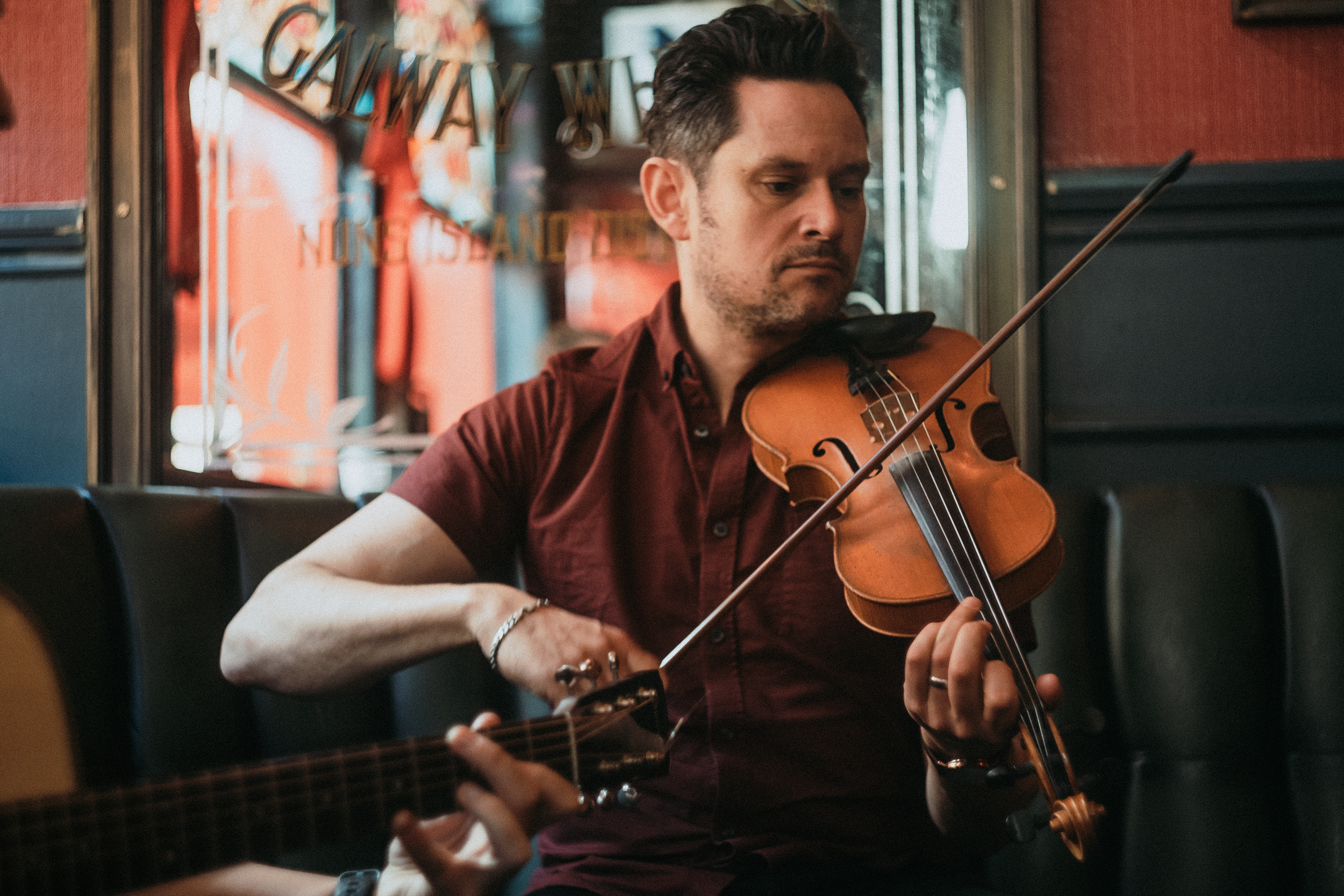 A man with dark hair and light skin playing a violin in an indoor setting, with a second person playing guitar partially visible in the foreground, behind a window with a decorative banner.