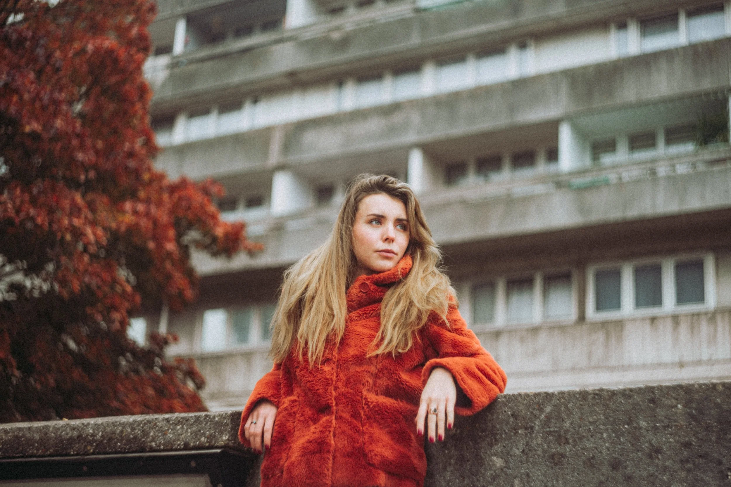 Young woman with long blonde hair leaning on a stone railing outdoors, wearing a red furry coat, with a multi-story concrete building and red autumn tree in the background.