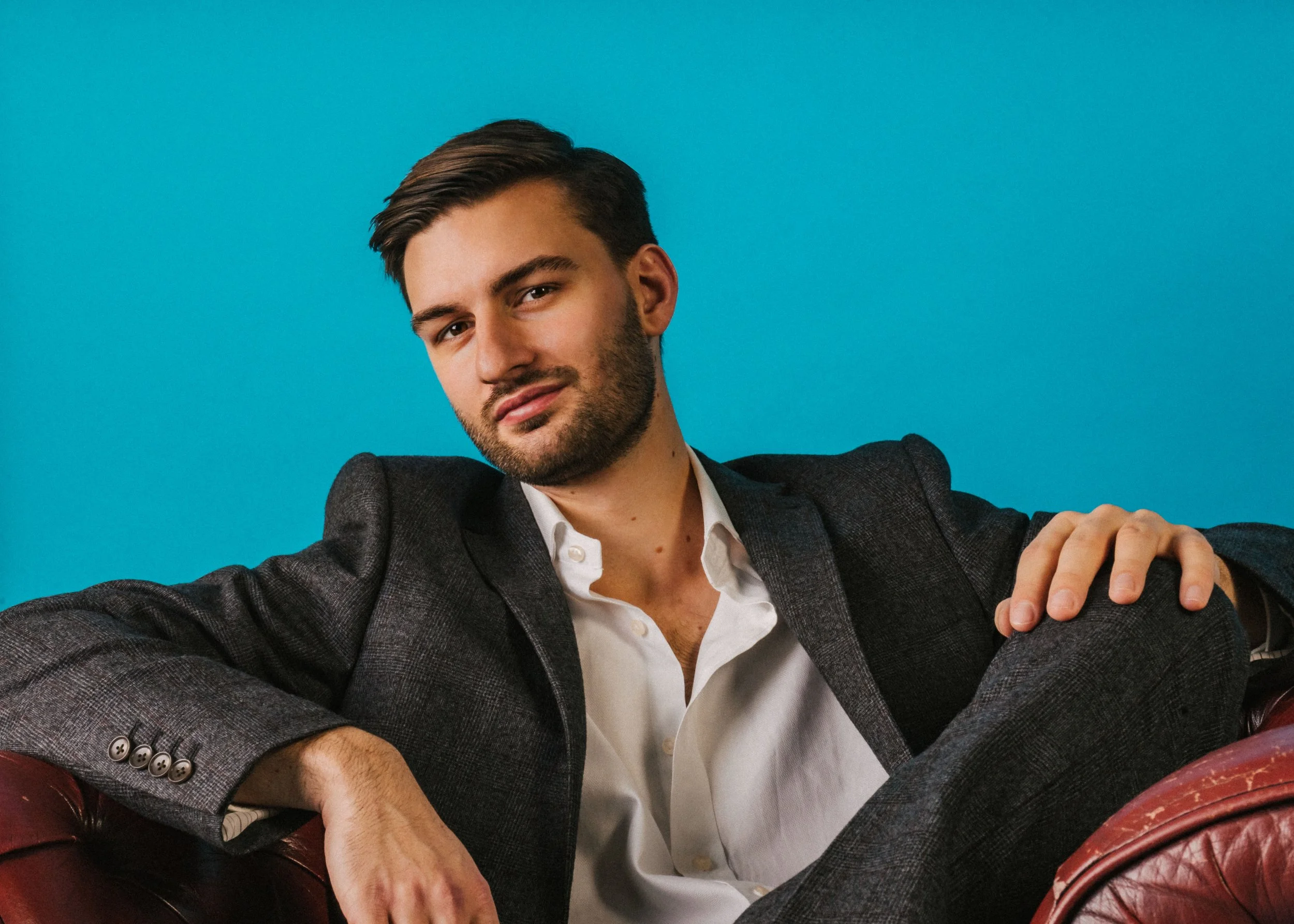 A man with dark hair and a beard wearing a gray blazer and white shirt, sitting on a red leather chair against a bright blue background.