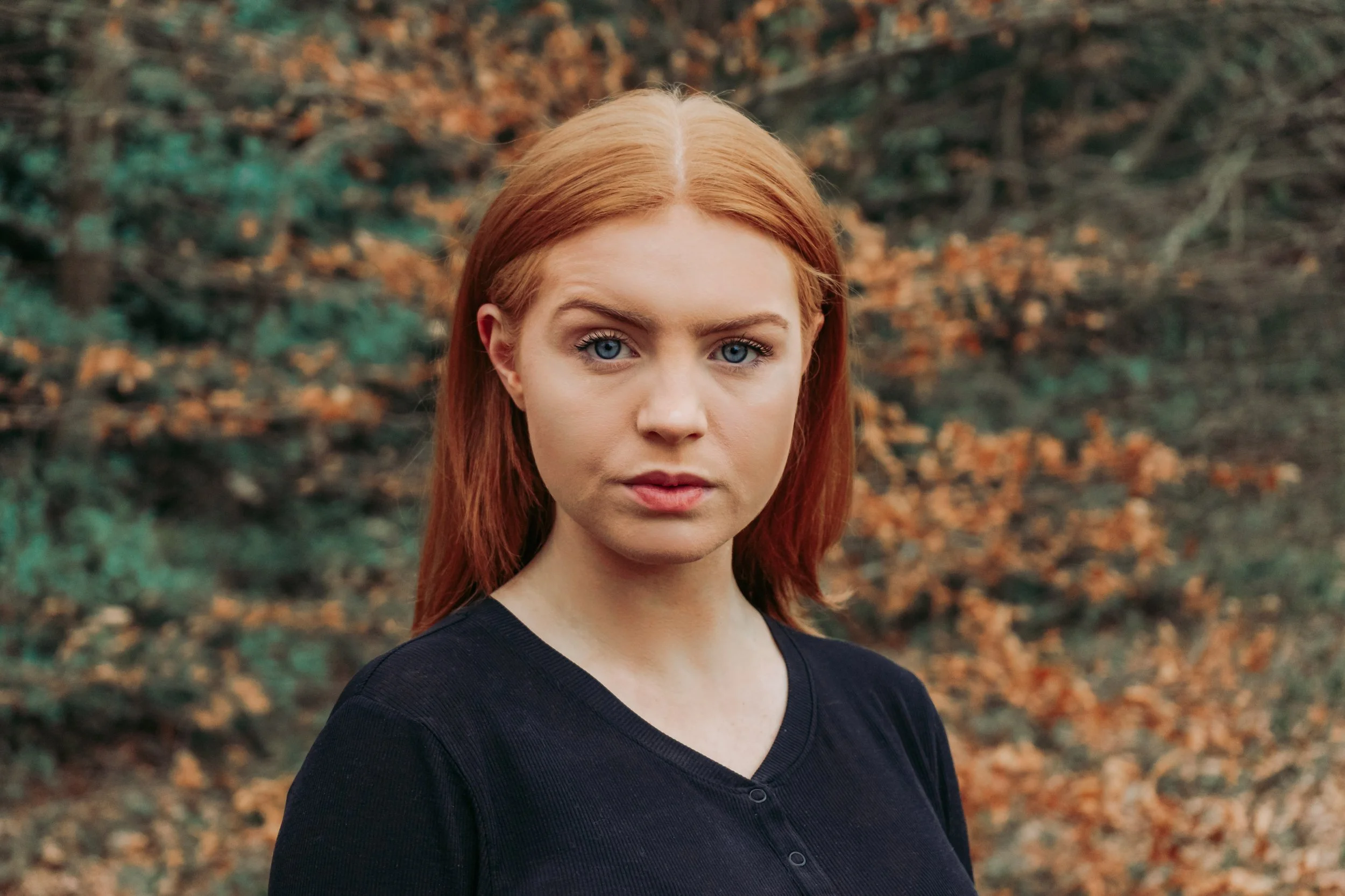 A young woman with red hair and blue eyes looking directly at the camera, outdoors with autumn-colored leaves in the background.