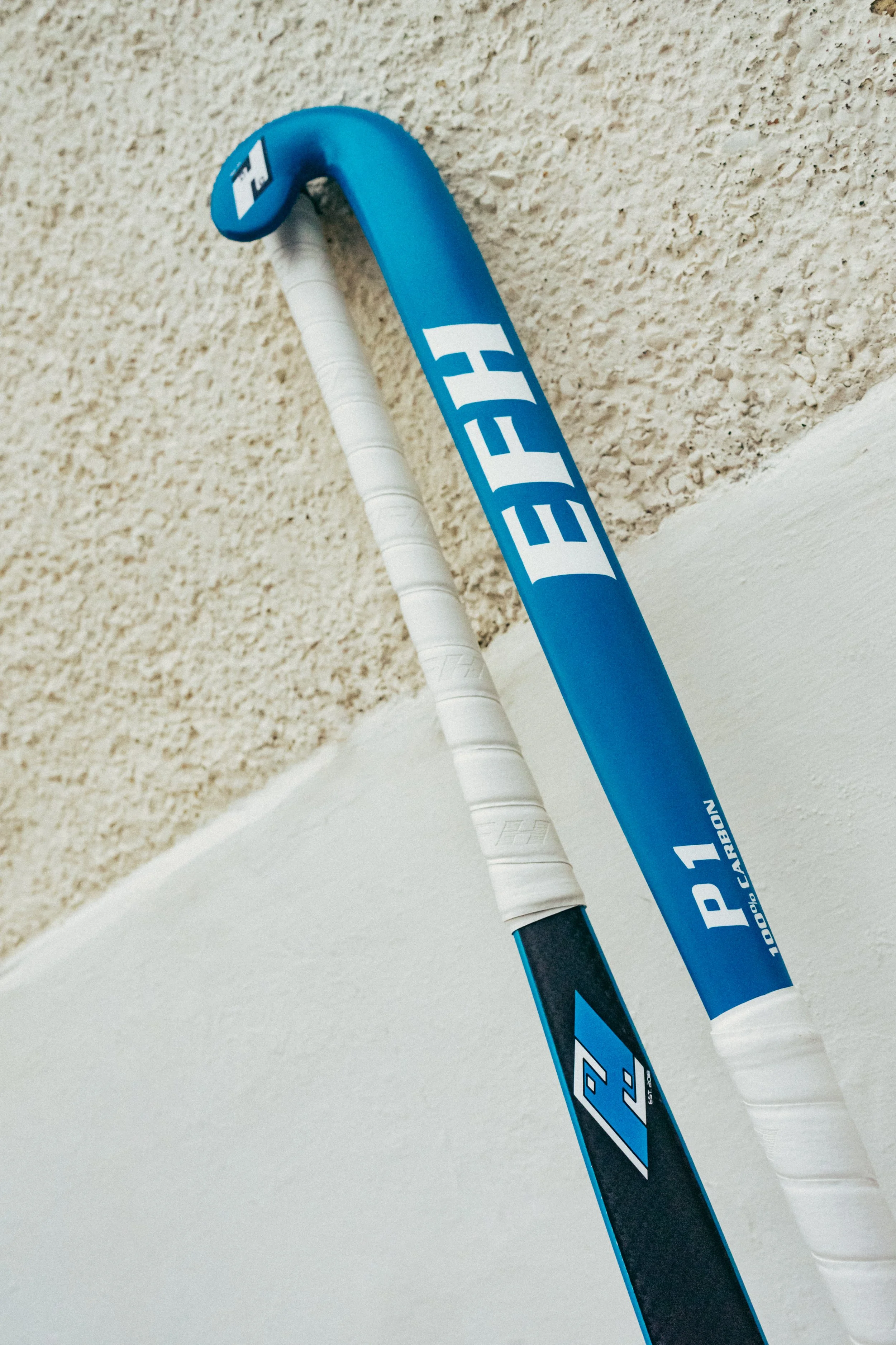 Close-up of a blue and white hockey stick, embedded in a wall, with part of a textured beige wall visible in the background.