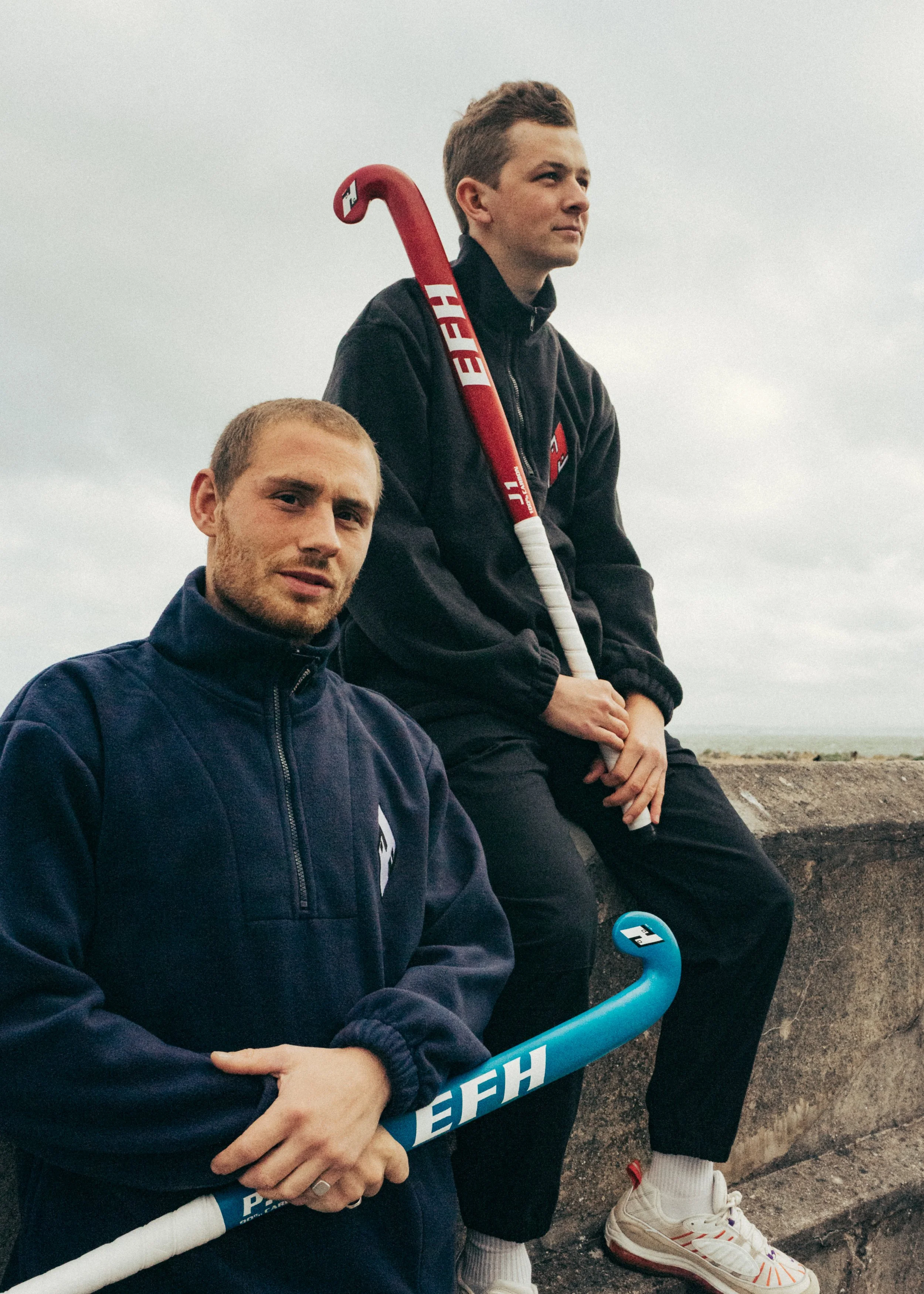 Two young men are sitting outdoors with a cloudy sky background, one holding a blue hockey stick with white lettering and the other sitting next to him.