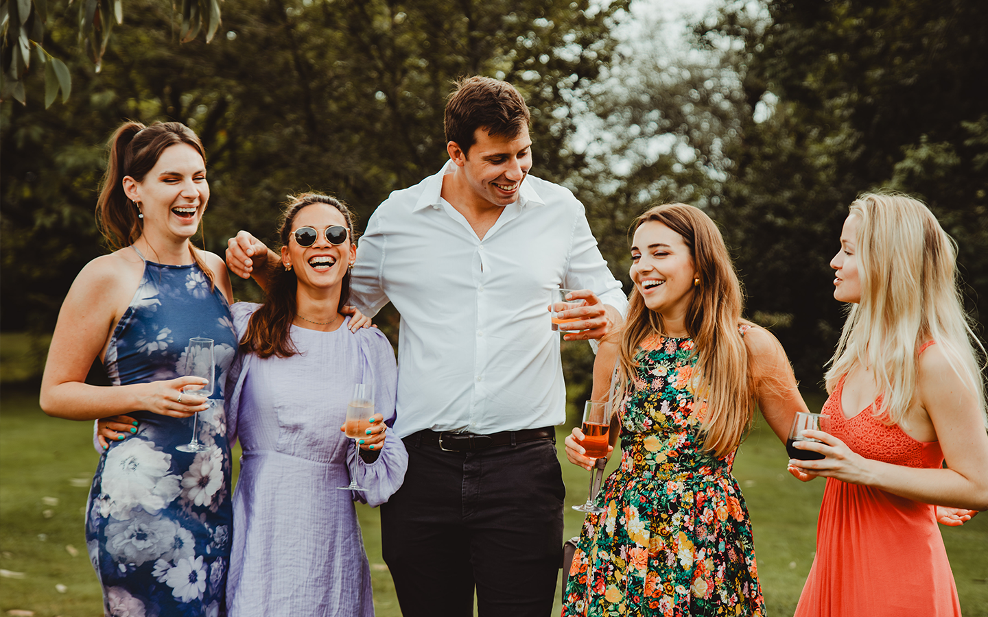 A group of six friends enjoying an outdoor gathering, smiling, and holding drinks near a lake with trees in the background.