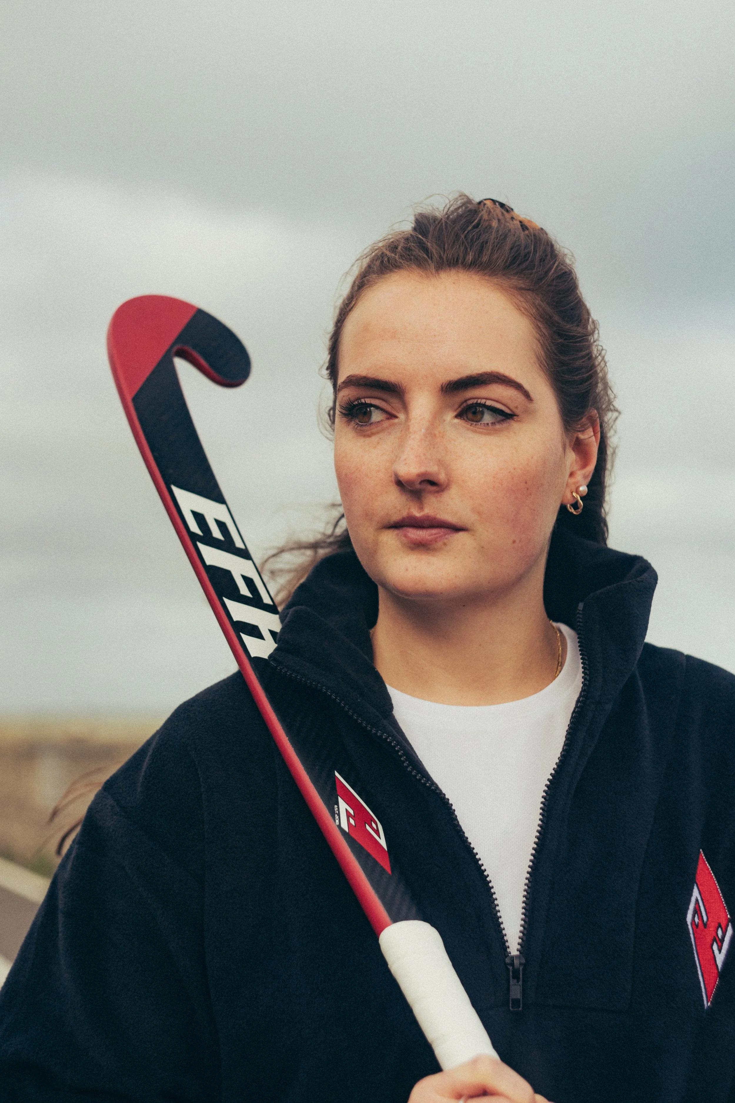 A young woman with brown hair pulled back, wearing earrings, a white t-shirt, and a black jacket, holding a hockey stick outdoors.