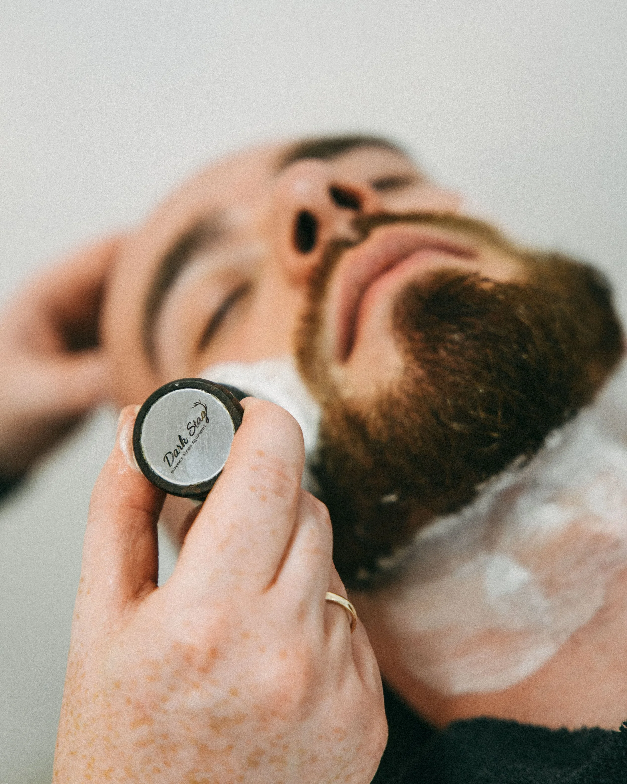 Close-up of a person with freckles applying face cream to their face using a small container labeled 'Dark Shade' while lying on a towel.