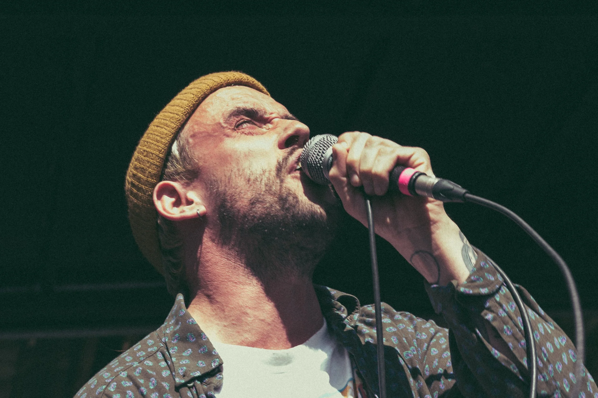 A man singing into a microphone, wearing a brown beanie and patterned shirt, with a concentrated expression.