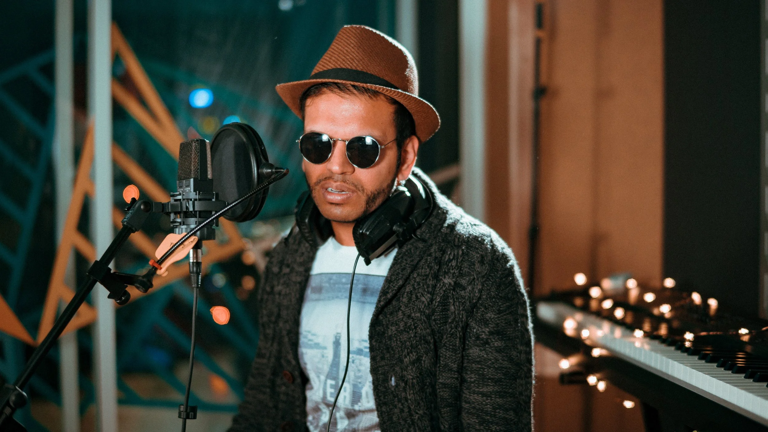 A man wearing a brown fedora, sunglasses, and a dark gray coat singing into a microphone in a recording studio with dim lighting and a piano in the background.