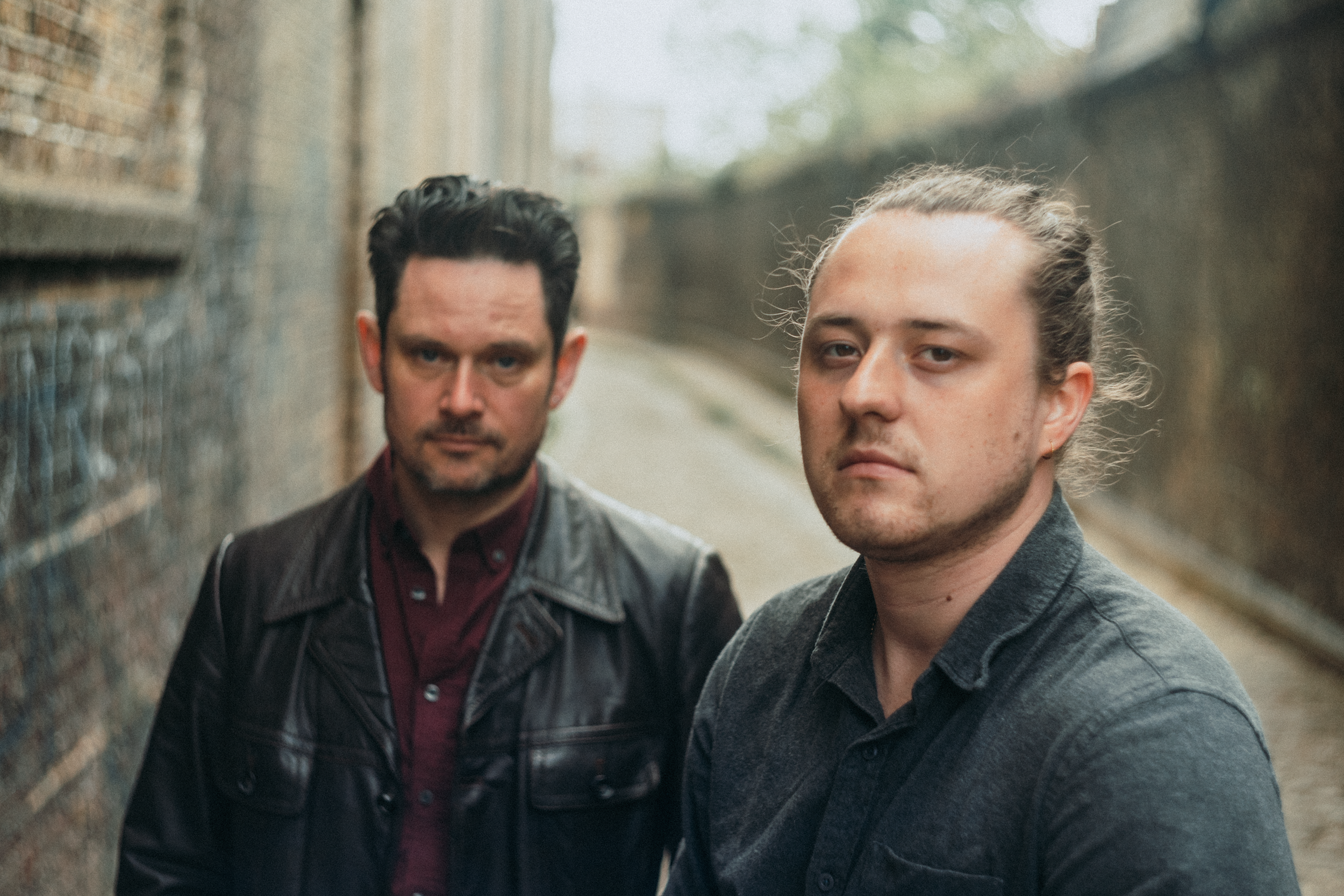 Two men with serious expressions standing outdoors in front of a brick wall with a blurred background.
