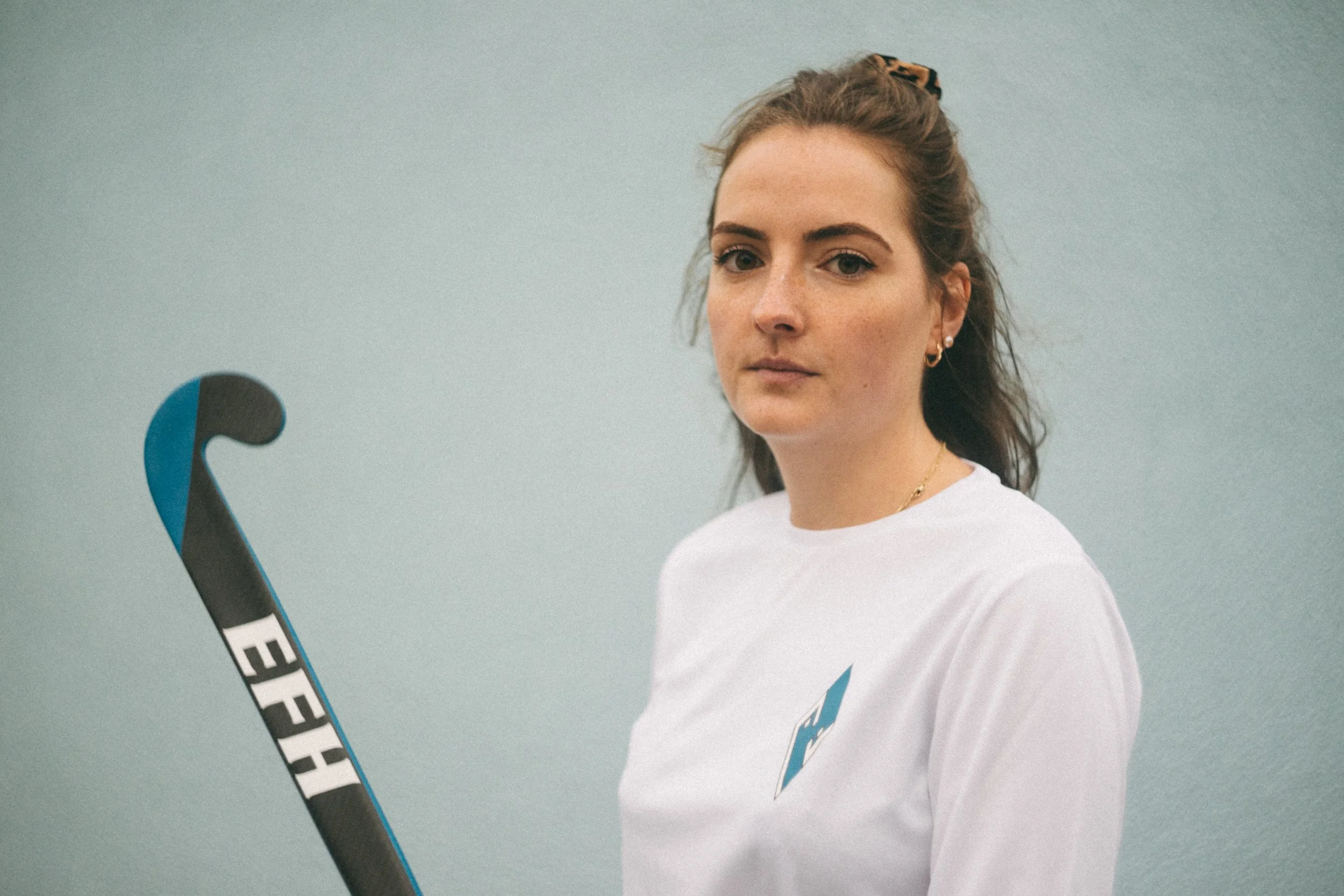 A woman with light skin and brown hair tied with a scrunchie, wearing a white shirt and hoop earrings, posing indoors against a plain light gray wall, with a tennis racquet in the foreground.