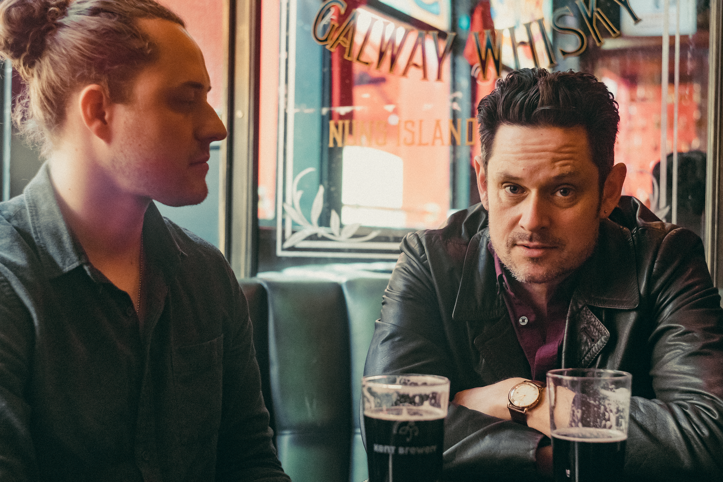 A woman with her hair tied up and a man with dark hair, both sitting at a bar table in a pub or restaurant, engaging in conversation. There are glasses of dark beer on the table. The background shows a window with a sign that reads 'GALA WAY WISH' an