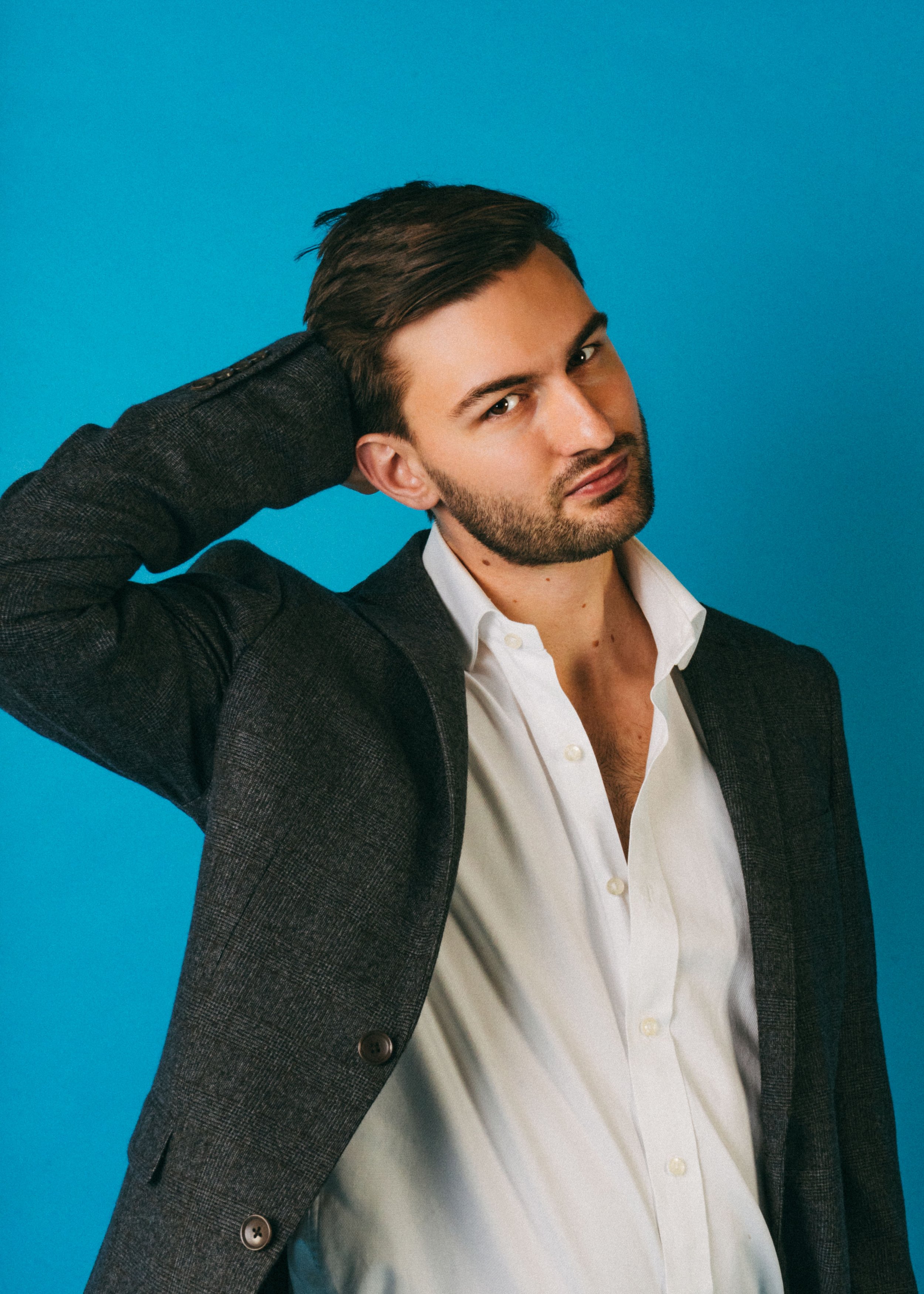 A young man with dark hair and a beard is posing with his left arm raised behind his head, against a bright blue background, wearing a white shirt and a dark blazer.