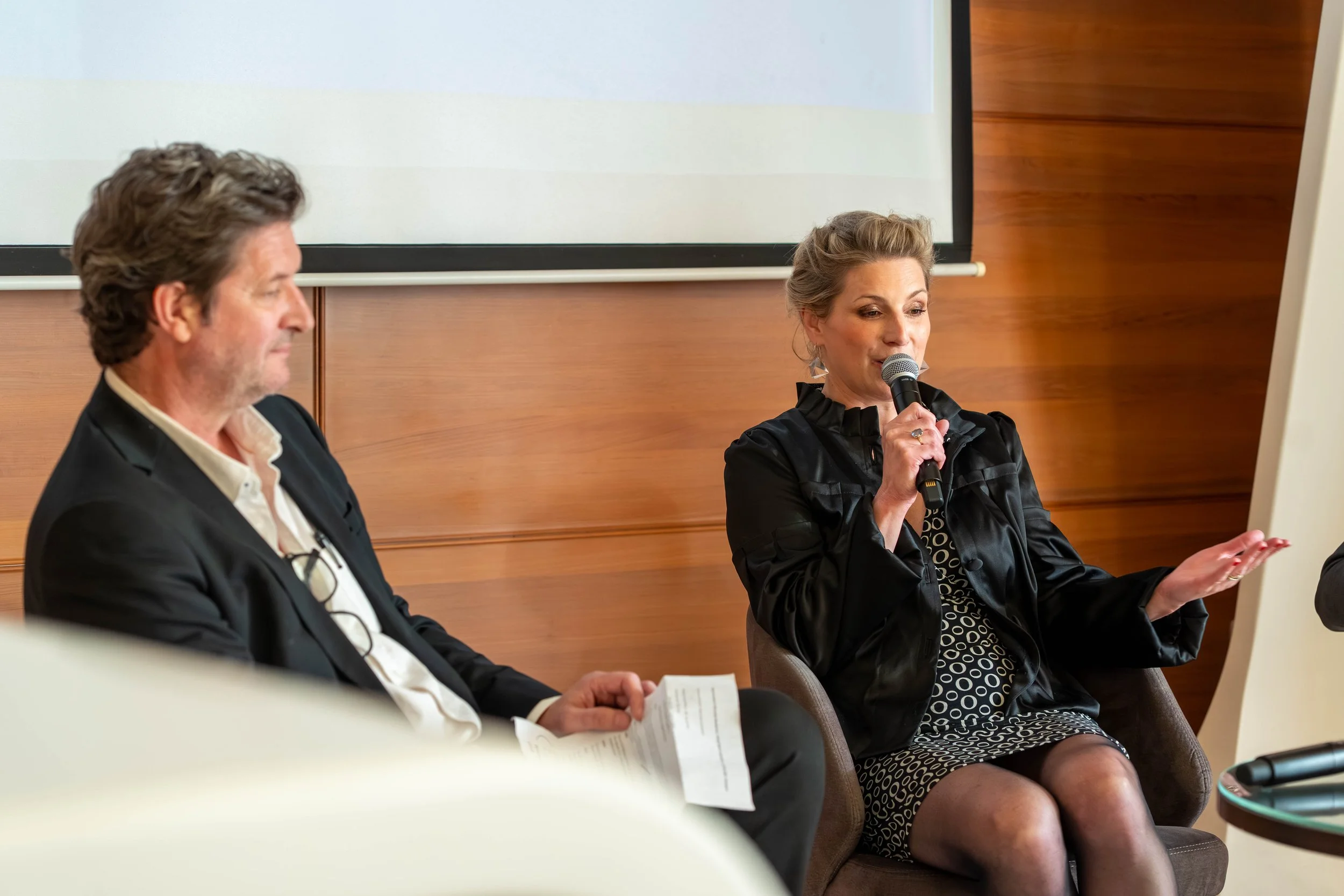 A woman speaking into a microphone while sitting in a panel discussion, with a man sitting to her left holding papers, in a conference room with wood-paneled walls.