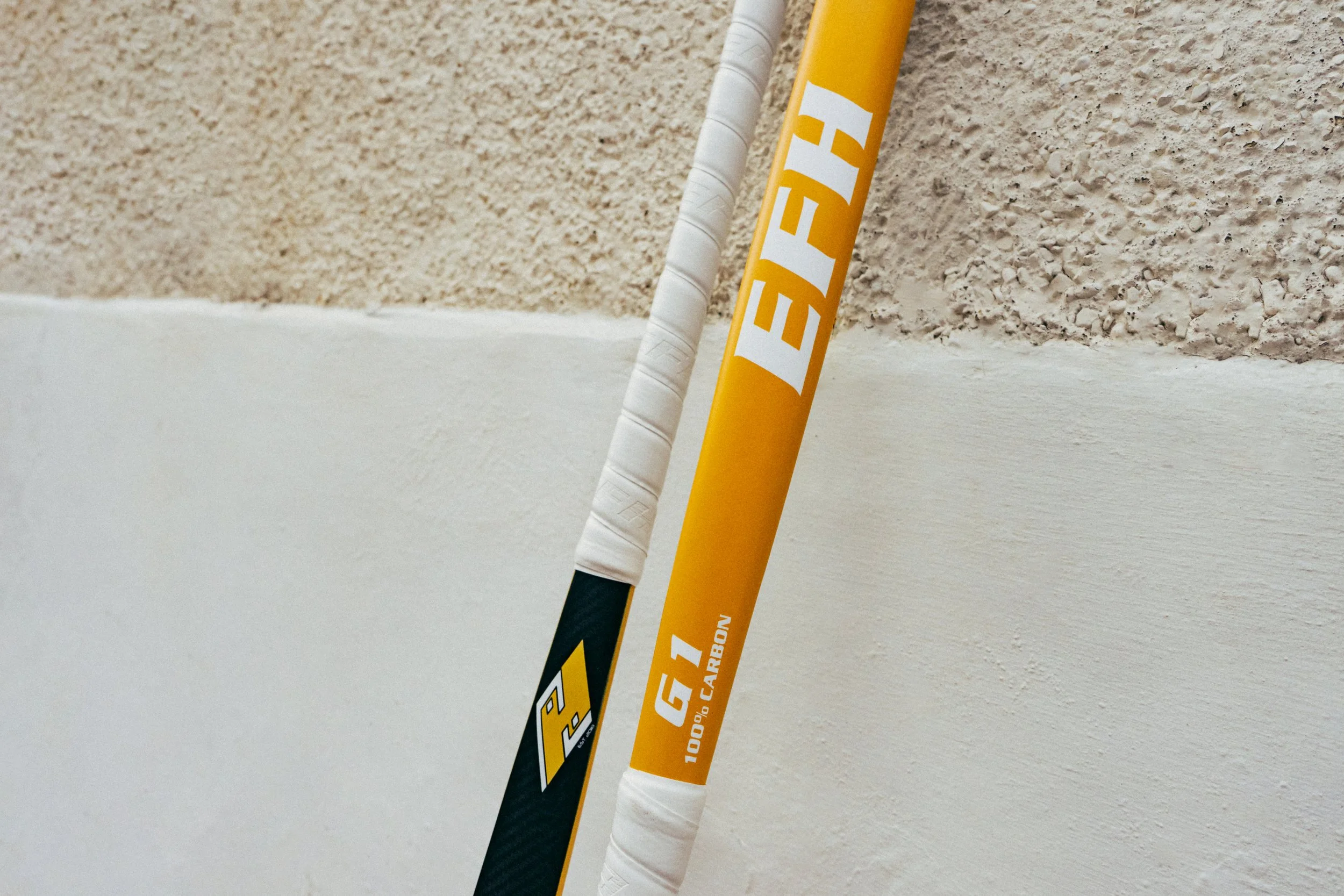 Close-up of two hockey sticks, one yellow and one black, leaning against a textured wall with a smooth white surface at the bottom.