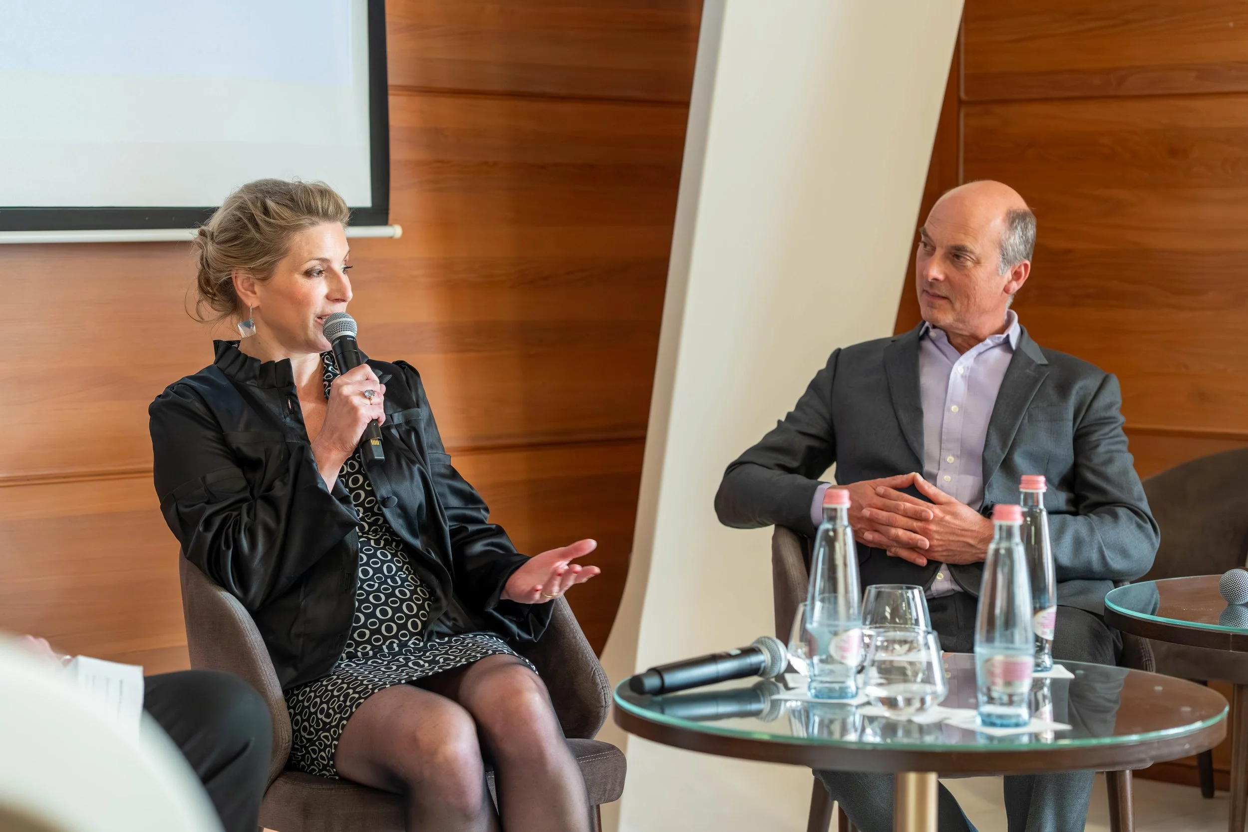 A woman holding a microphone and speaking while seated at a panel discussion, with a man listening attentively. They are in a room with wooden walls, and there are bottles of water on the table in front of them.