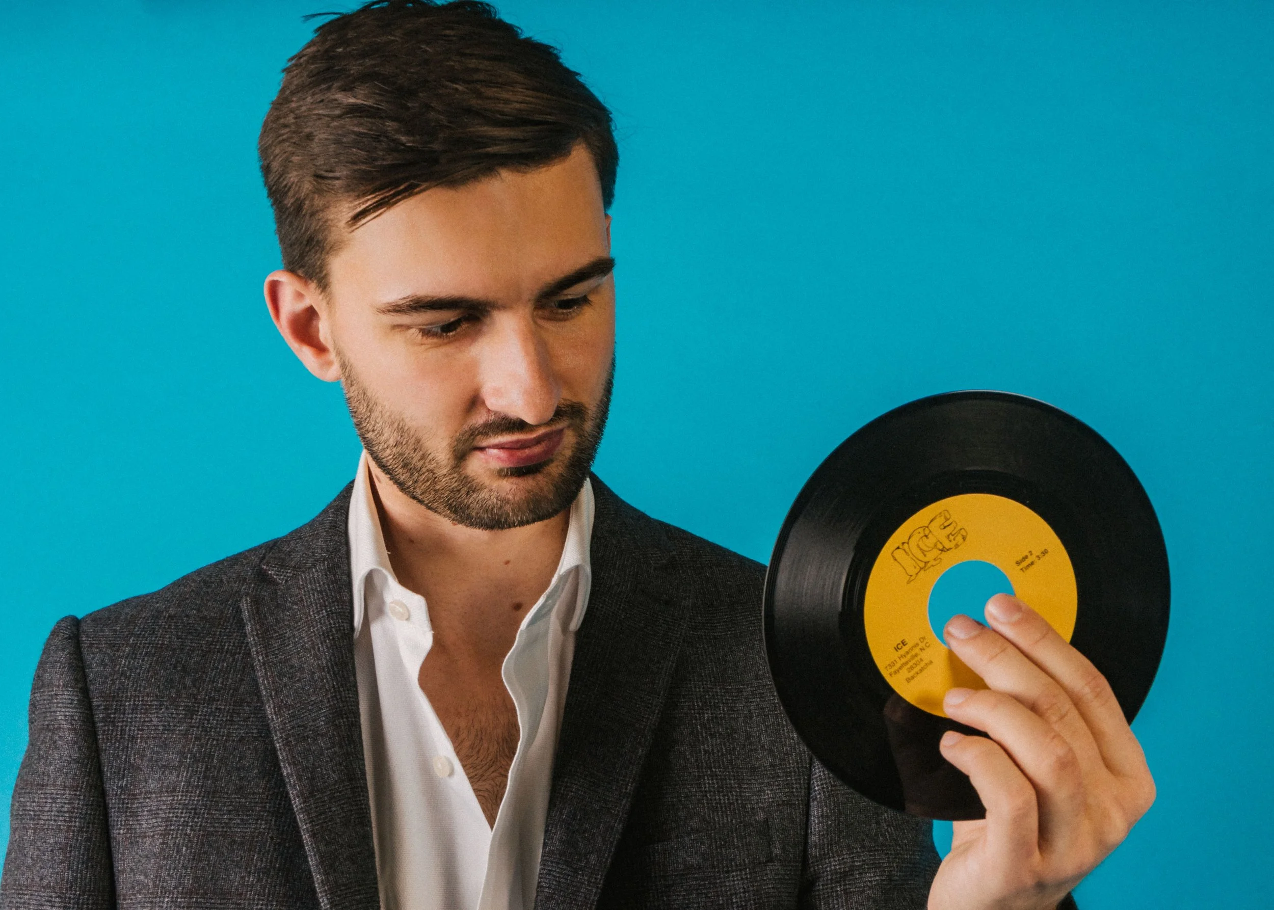 A young man with dark hair and a beard holds a vintage black vinyl record with a yellow and blue label against a bright blue background.