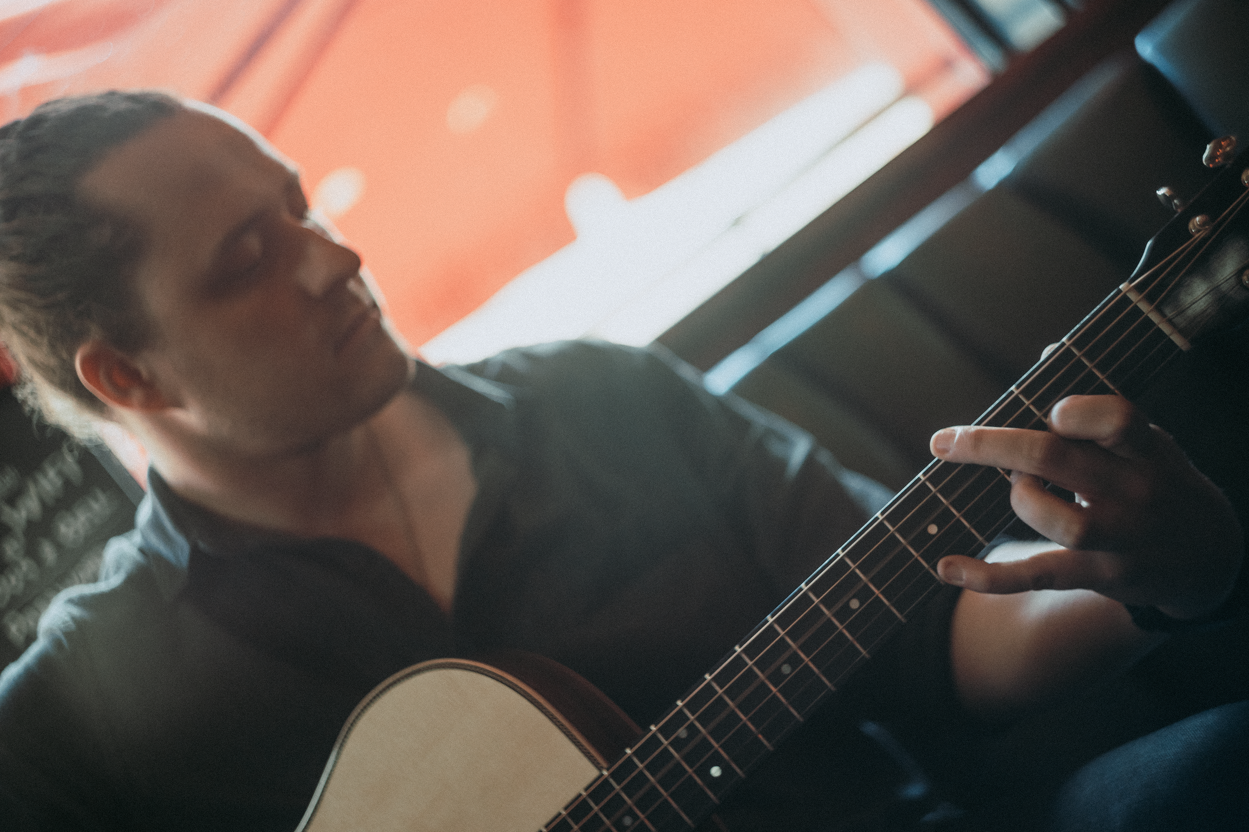 Person playing an acoustic guitar indoors with orange walls and a window in the background.