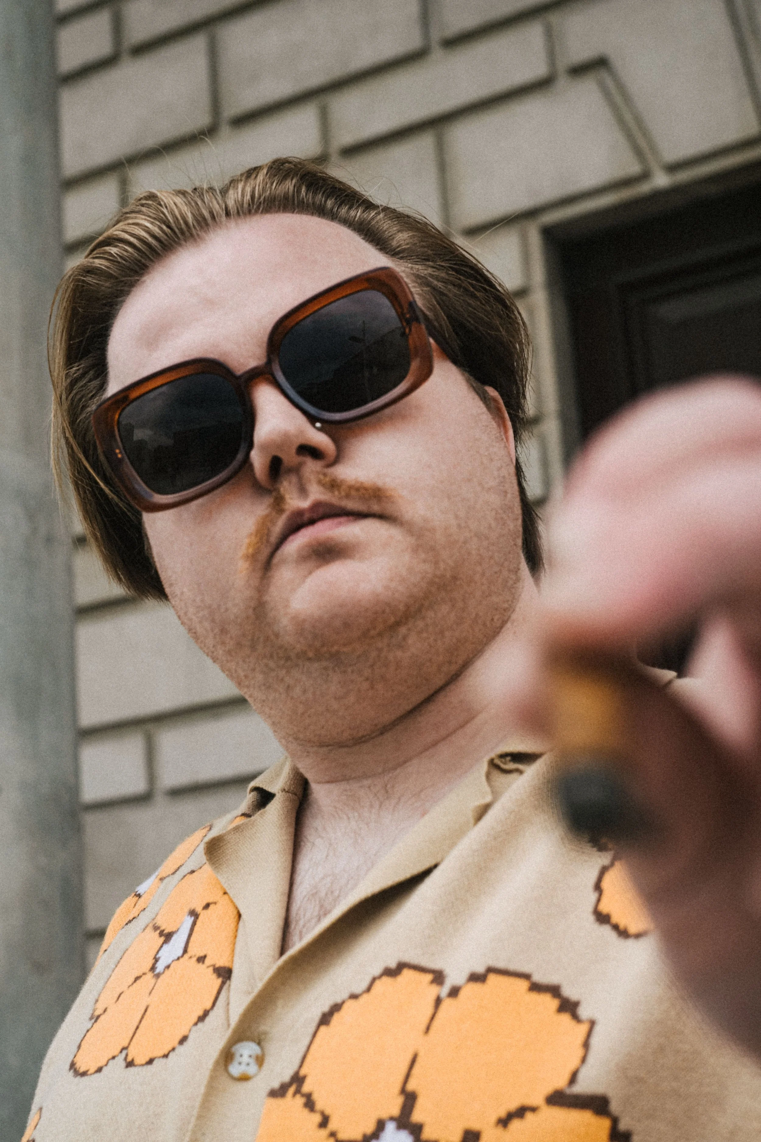 Close-up of a man wearing large sunglasses and a beige shirt with an orange pattern, standing outside with a brick wall in the background.