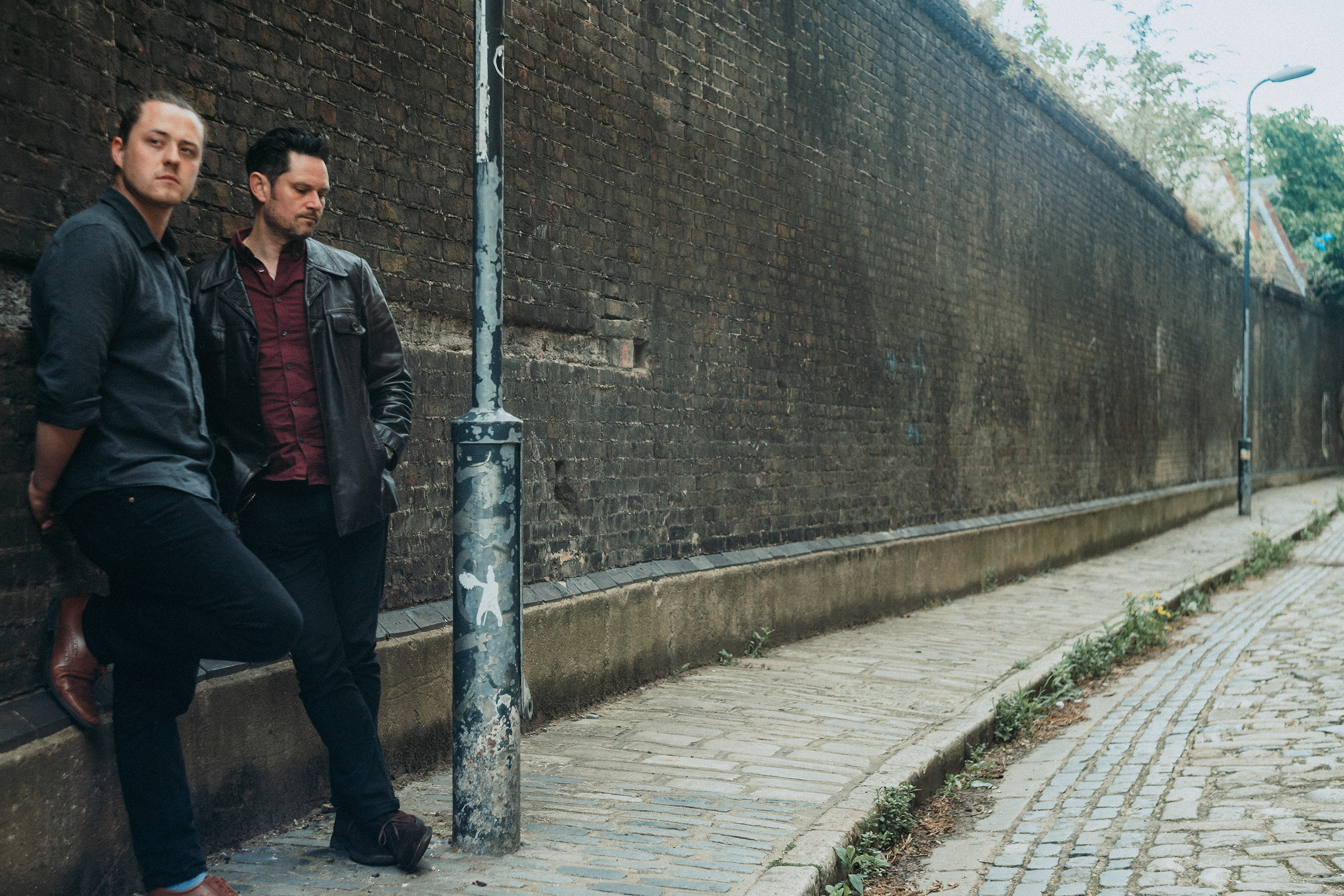 Two men with dark hair, wearing dark clothing, standing against a brick wall on a sidewalk. One man has a leather jacket and the other a button-up shirt. They appear to be deep in thought or waiting.