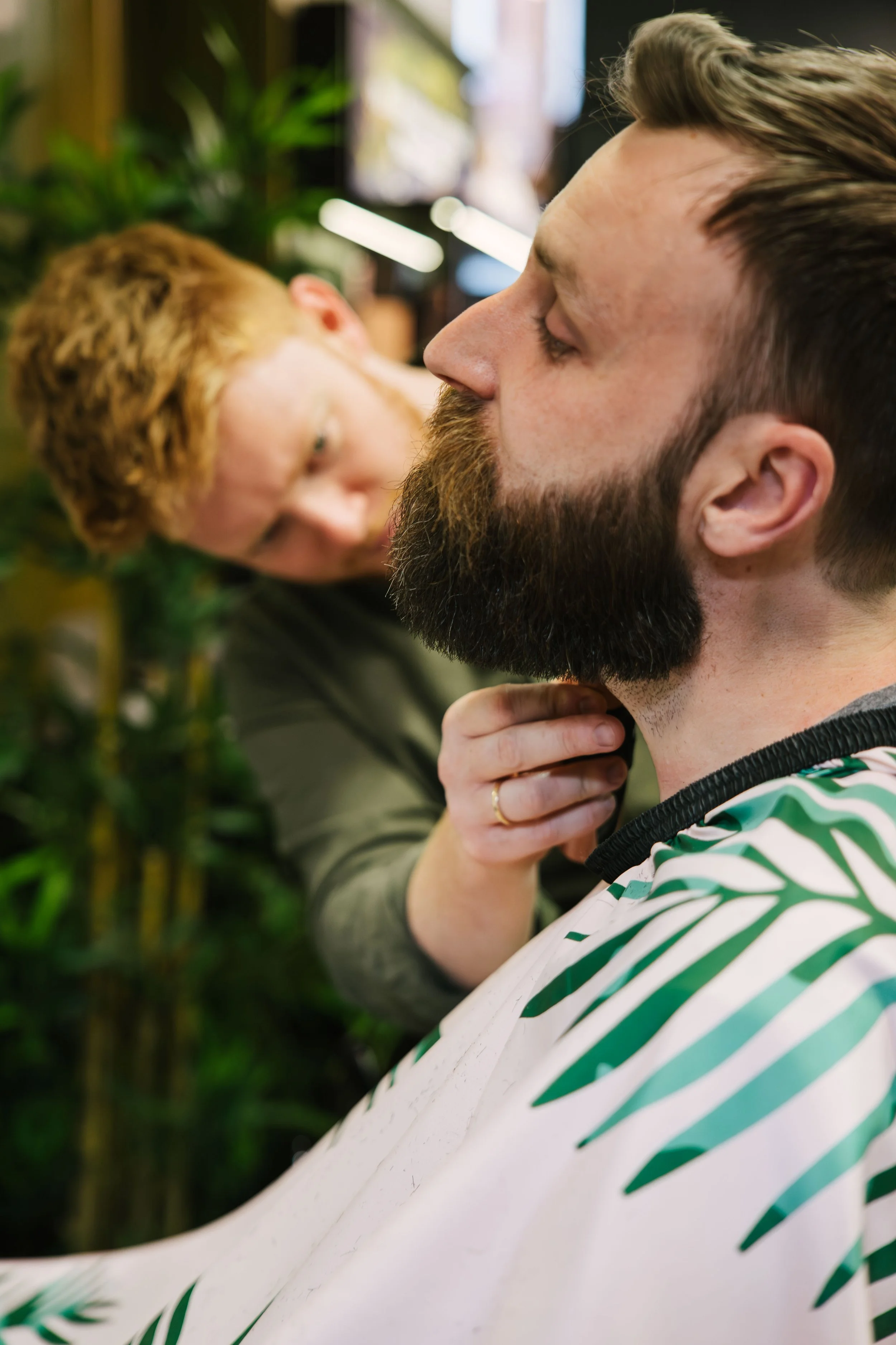 A man with a beard and short hair wearing a green and white striped shirt sitting in a barber chair while a hairstylist or barber gently touches his chin, in a salon or barbershop with plants and blurred background.