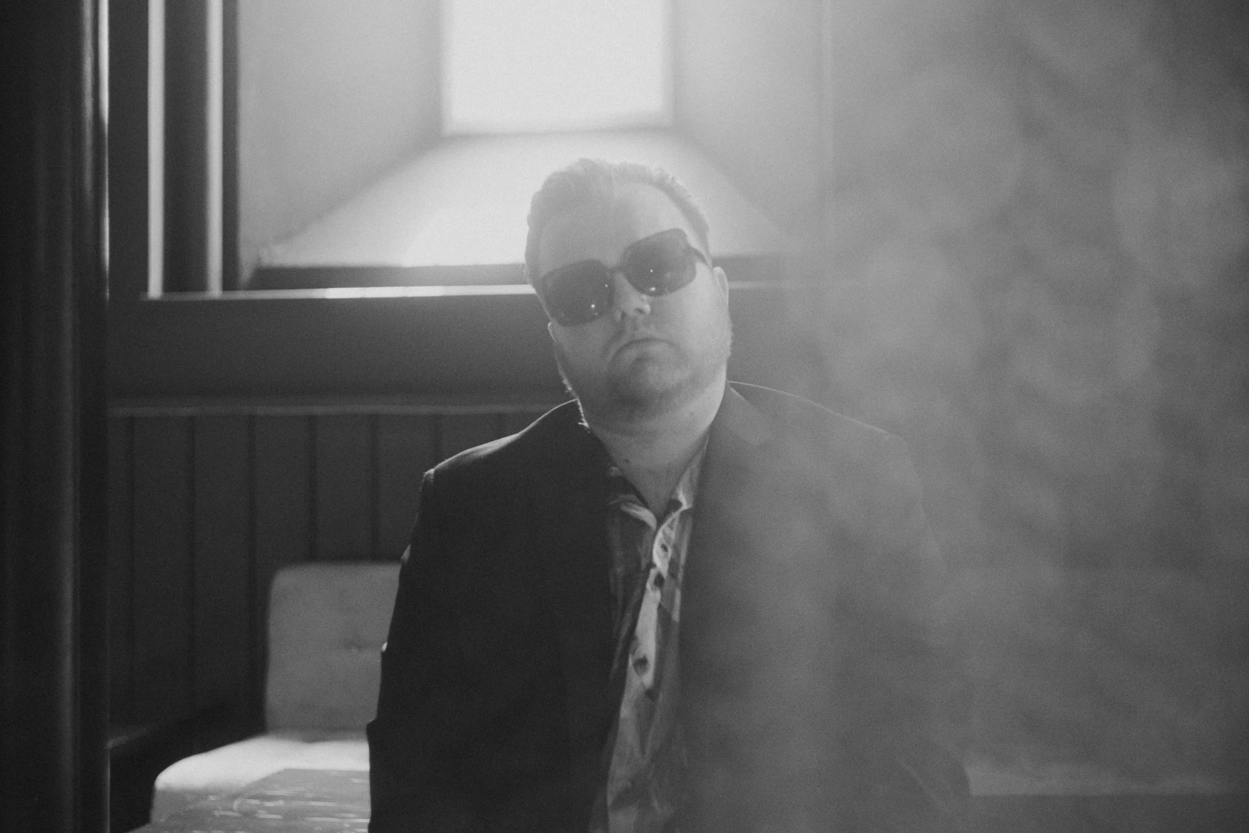 A black-and-white photo of a man with short hair and a beard, wearing sunglasses and a dark jacket, in a room with a lamp and wooden paneling. The photo is taken through a reflection or a mirror with some haze or fog.