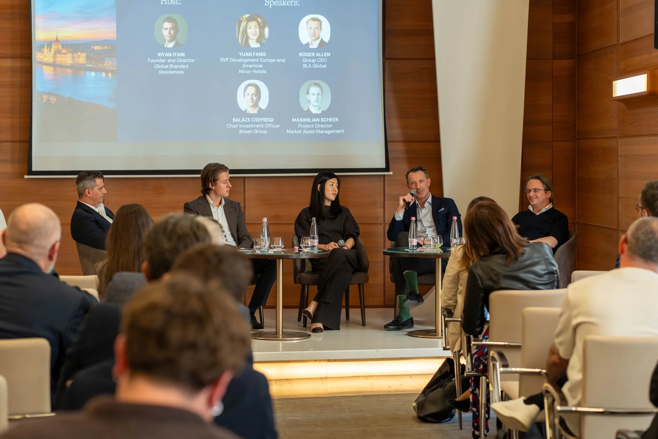 A panel of five people sitting on stage at a conference, with an audience in front. Behind them is a large screen displaying names and titles of the panelists, and a scenic image of a city with a river at sunset.