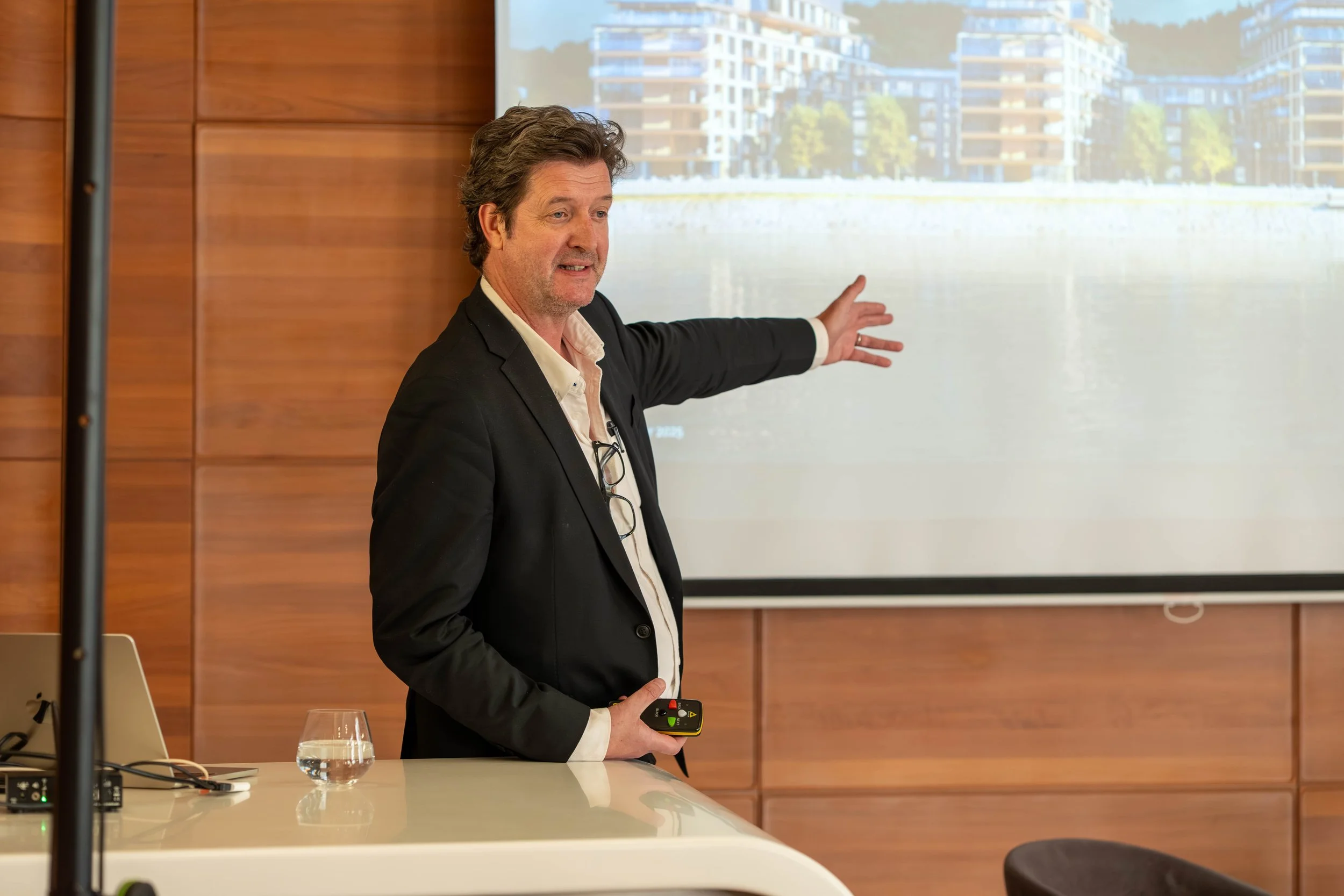 A man in a black suit giving a presentation, pointing at a projected image of modern buildings with trees in the background, in a conference room with wooden walls.