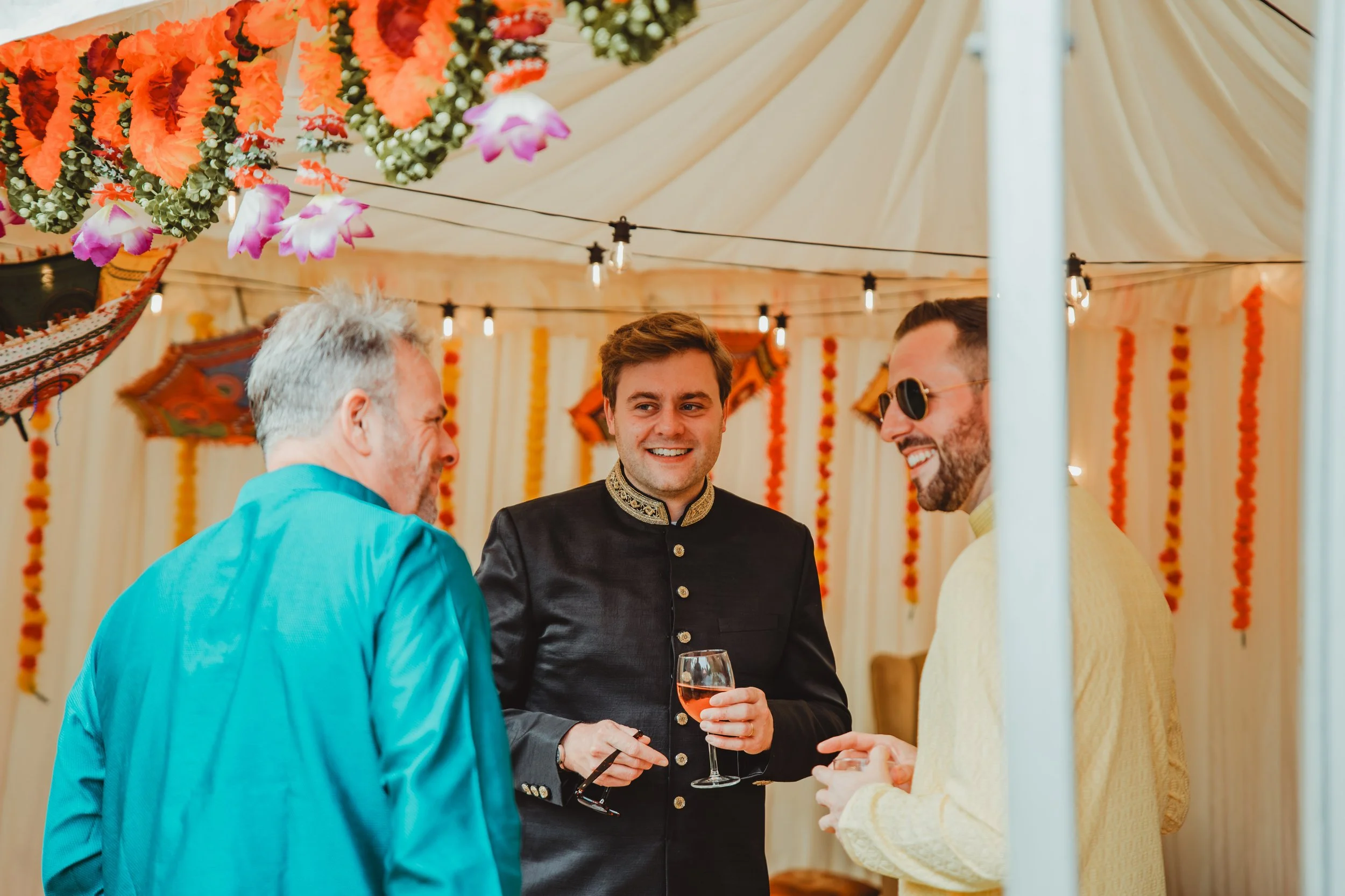 Three men are gathered in a decorated tent, smiling and engaging in conversation. One man holds a glass of rosé wine. The tent is decorated with orange flowers, green foliage, hanging lights, and colorful fabric walls.