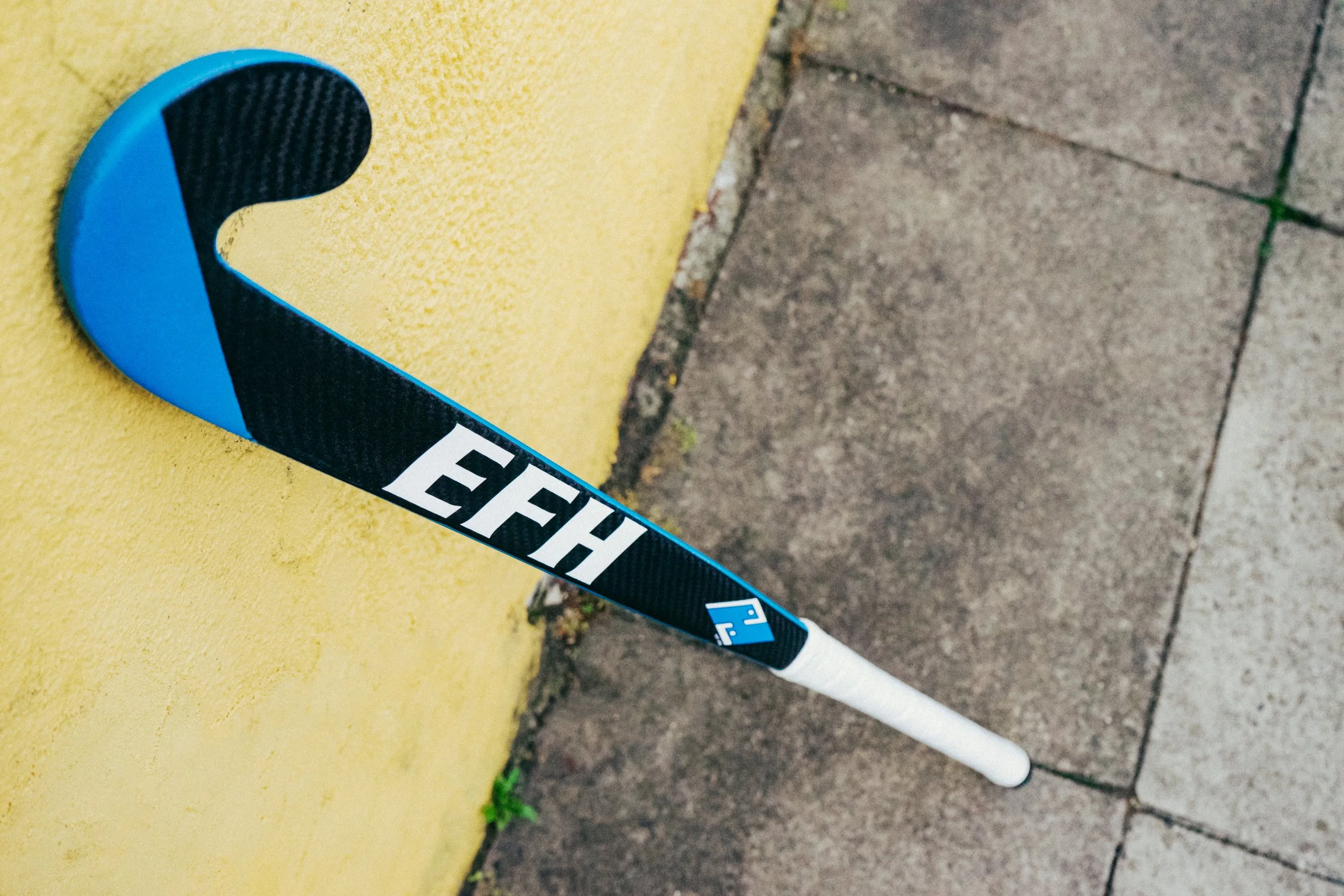 Close-up of a hockey stick resting on a yellow wall and concrete sidewalk.