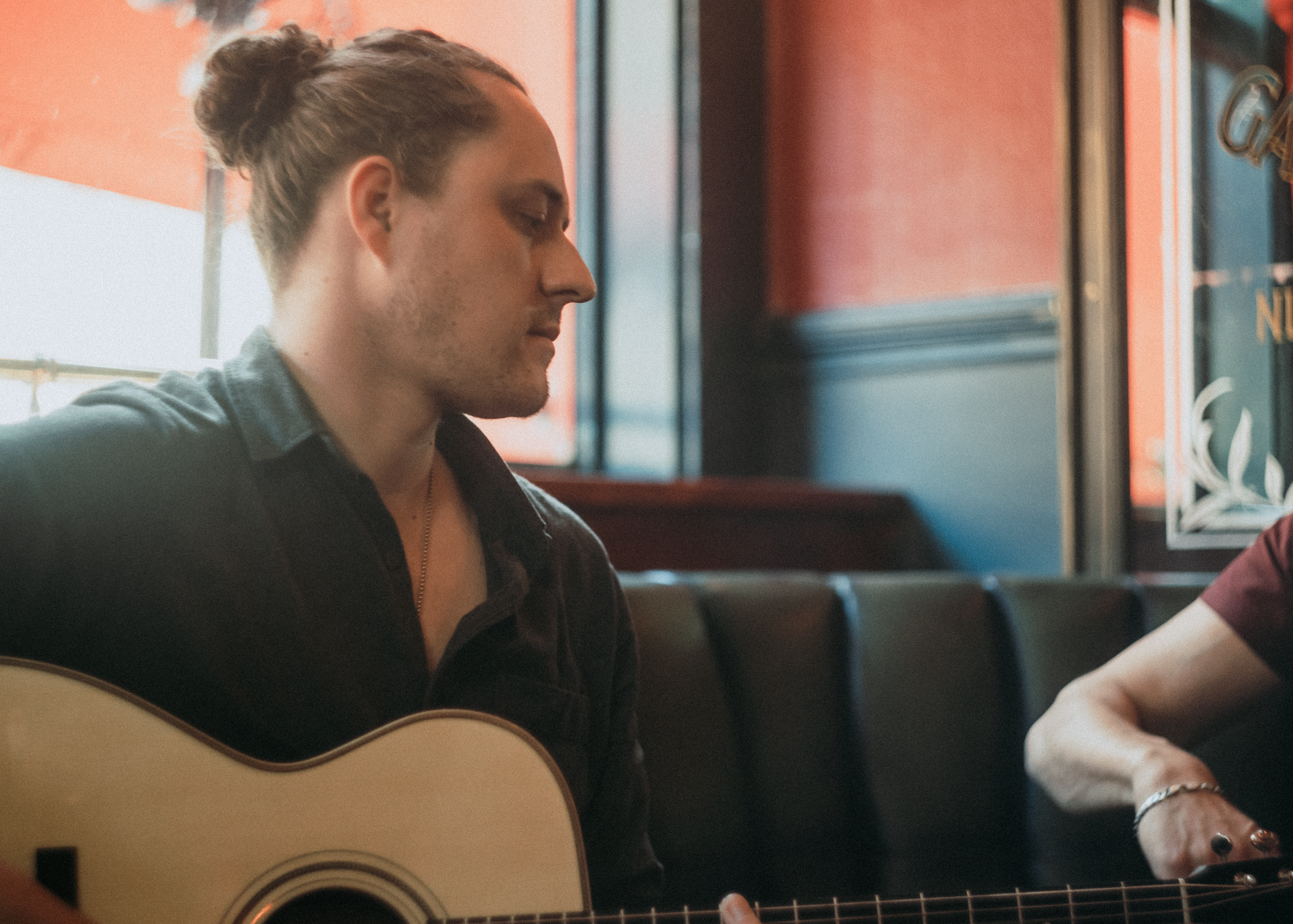 A person with dark hair tied in a bun playing an acoustic guitar, sitting in a warmly lit room near a window with a red wall behind.