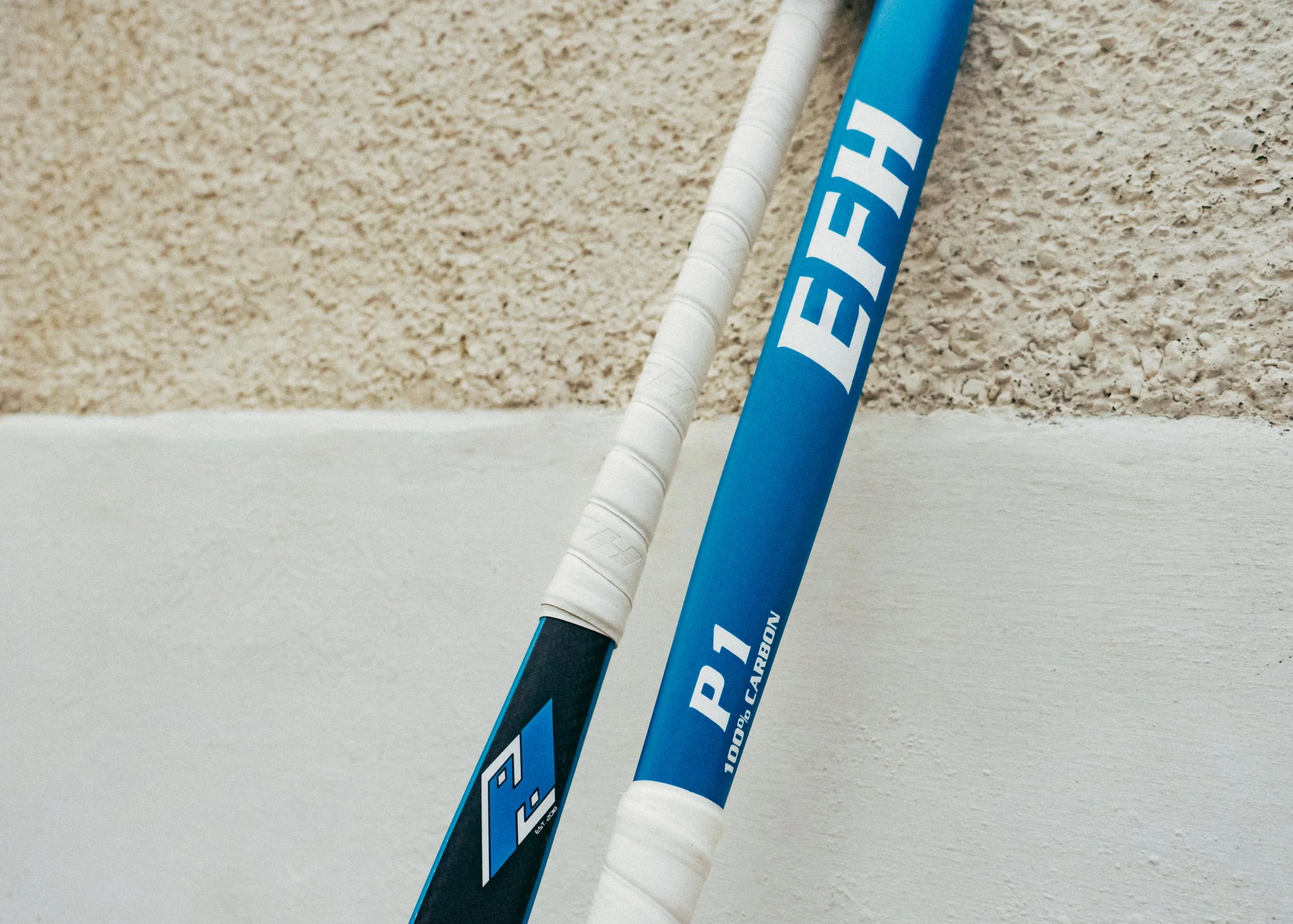 Close-up of blue and black hockey sticks with wrapped white grips, leaning against a beige textured wall.
