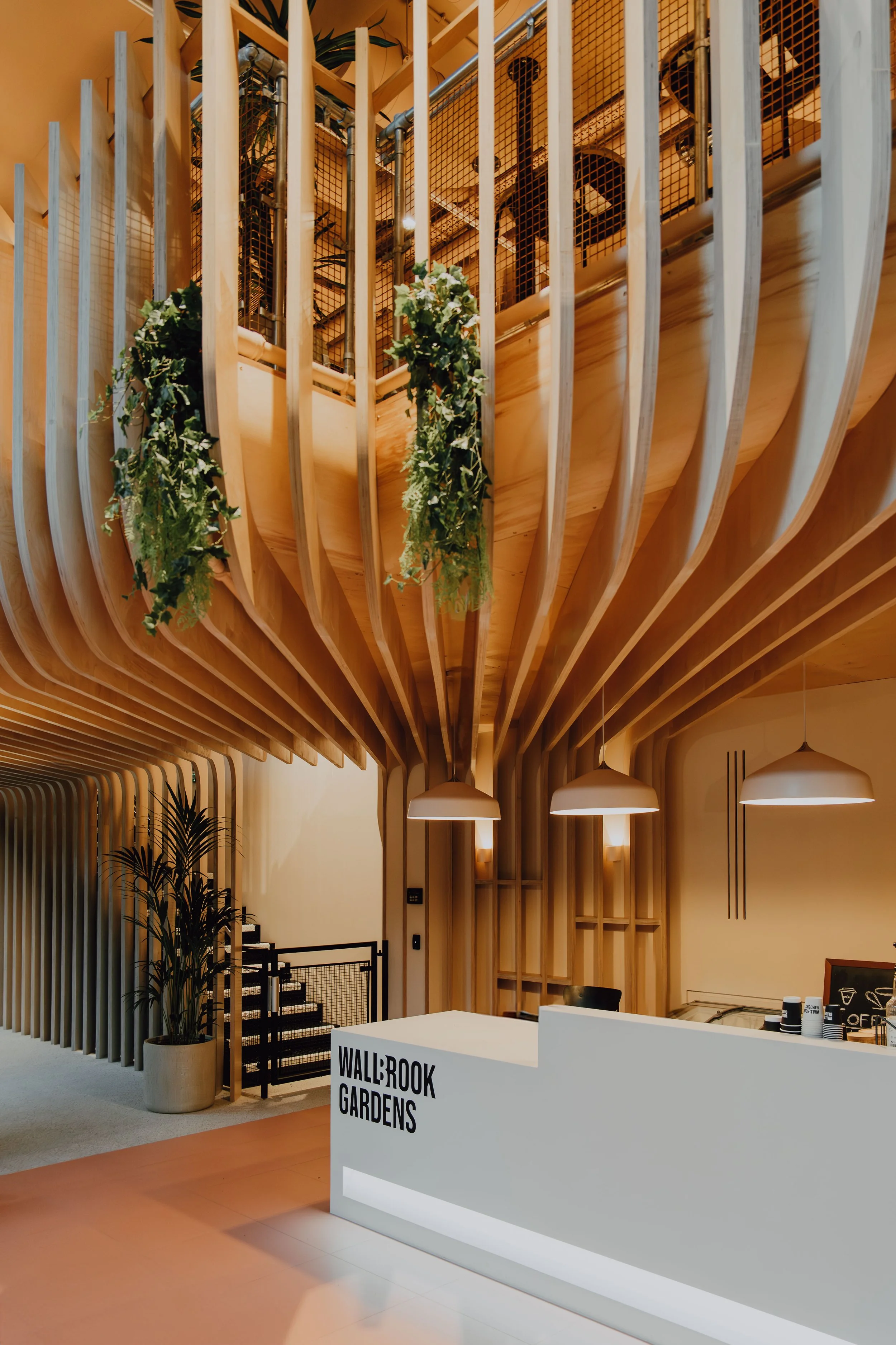 Interior of a modern building with wooden architectural elements and hanging lights. There is a reception counter with the words 'WALBROOK GARDENS' displayed on it, and greenery decor is visible, including plants and hanging foliage.